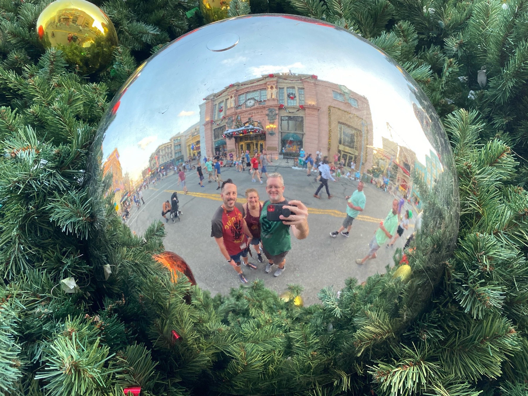 A group of three people (one female and two males) take a photo of their reflection in a giant, silver Christmas tree ornament with buildings in the background.