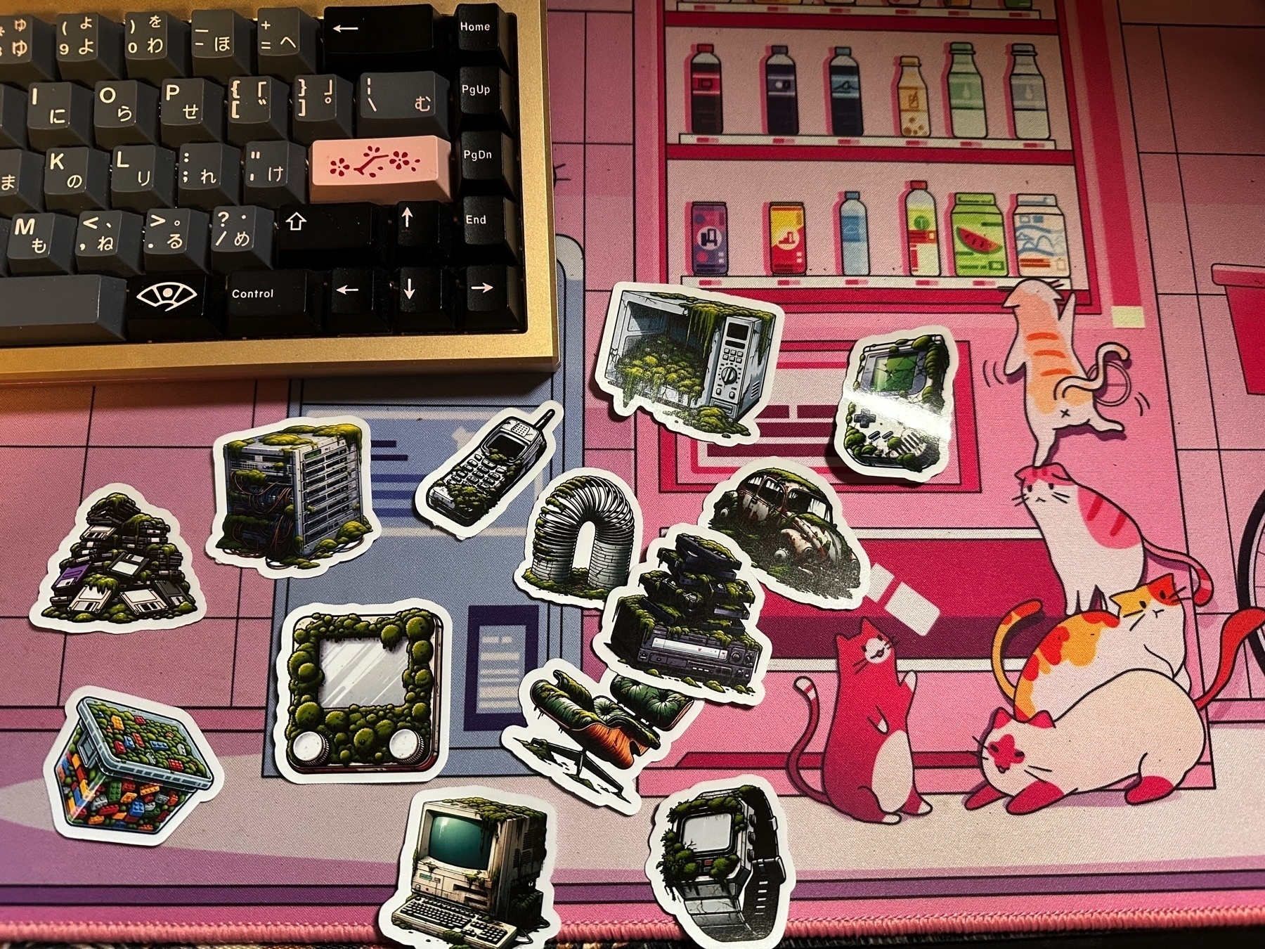 This image shows a variety of stickers scattered on a pink desk mat featuring a design with cartoon cats in a cafe setting. Among the stickers, there are illustrations of retro computers, computer parts, and olive-filled containers. On the left side, a mechanical keyboard with custom keycaps is partially visible. The scene conveys a playful and creative workspace vibe.