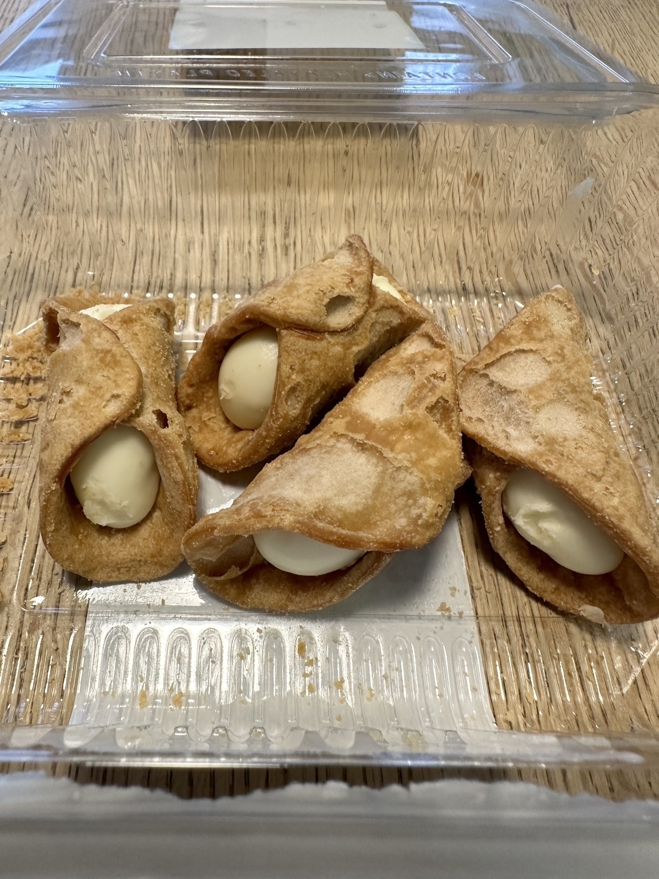 Four cannoli with a crisp, golden-brown shell, each filled with a generous amount of creamy white ricotta filling, are displayed in a clear plastic container on a wooden table. The container lid is open, revealing these classic Italian pastries in a ready-to-serve state.