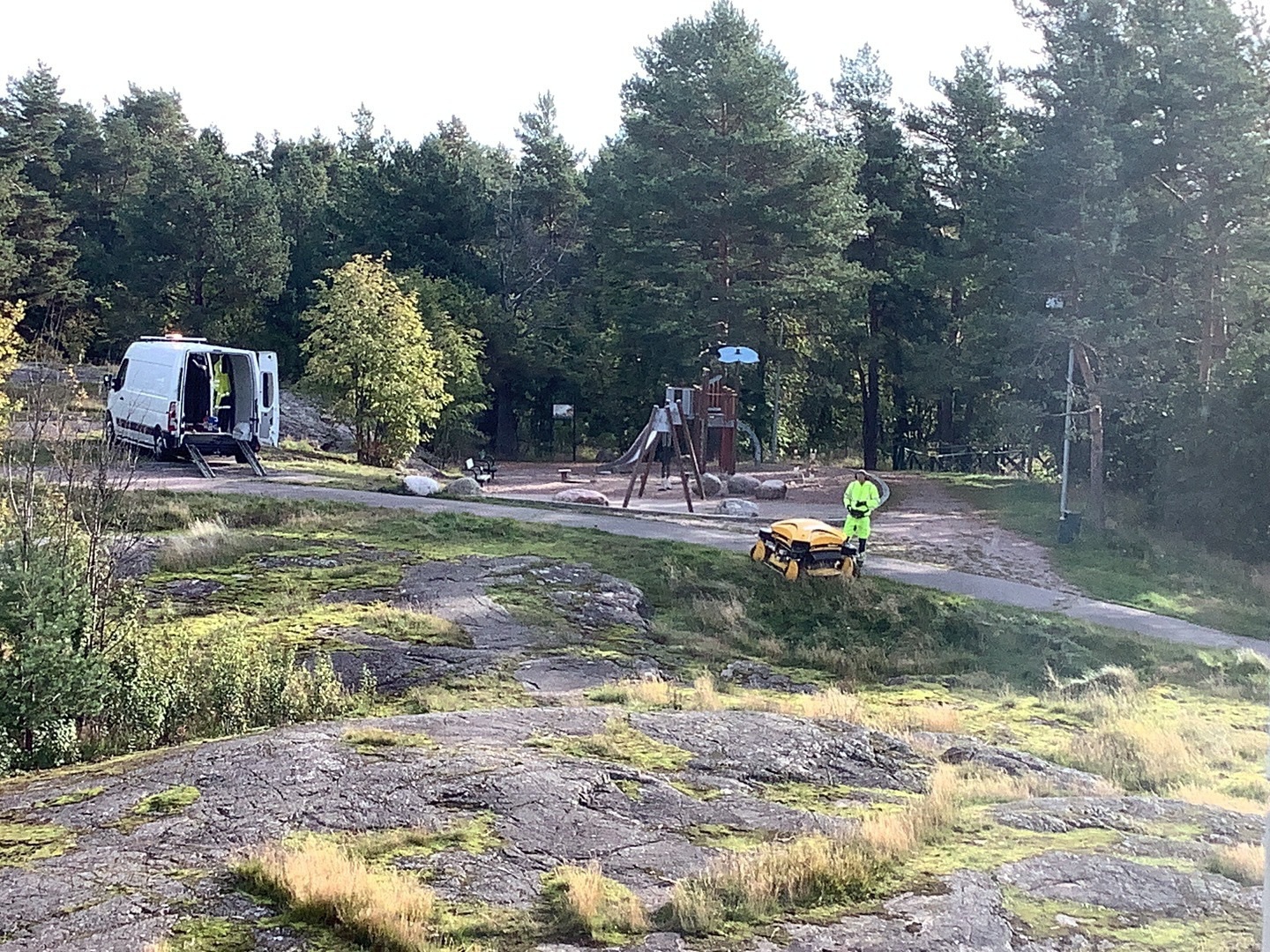 A park, with playground, rocks, a van and a robot lawn mower