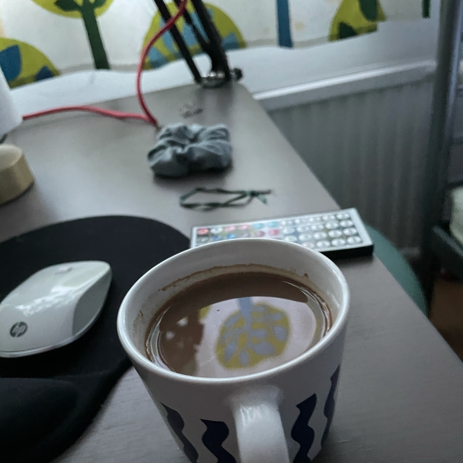 cup of coffee, a couple of light controller remotes and a hair crunchy on a desk corner