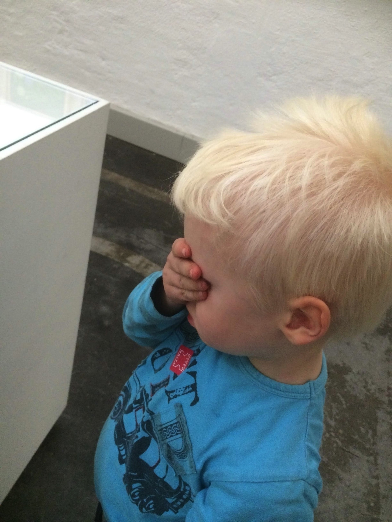 boy, three years old standing by a art piece blocking his eyes with a hand. looking unhappy