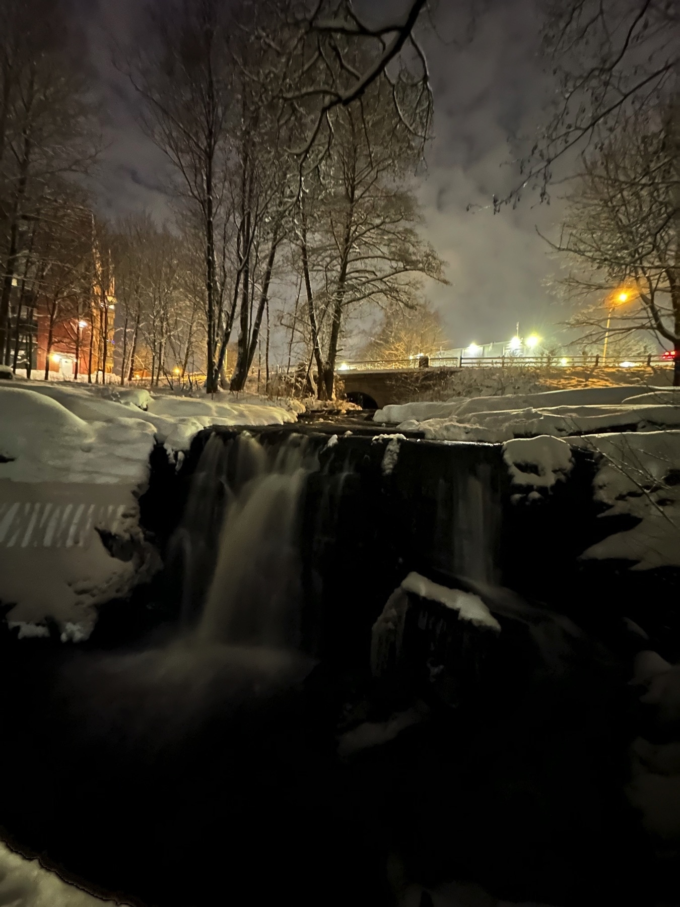 city lights shining behind a small waterfall surrounded by snow