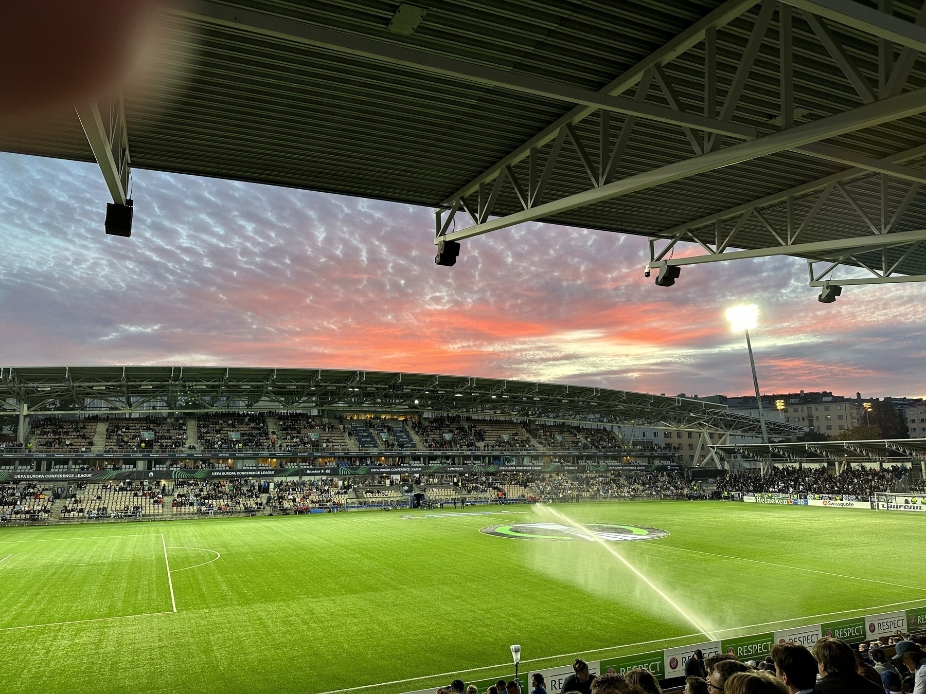 Football stadium, clouds reflecting the red of the setting sun. Bright green football pitch being sprayed with a stream of water from a jet