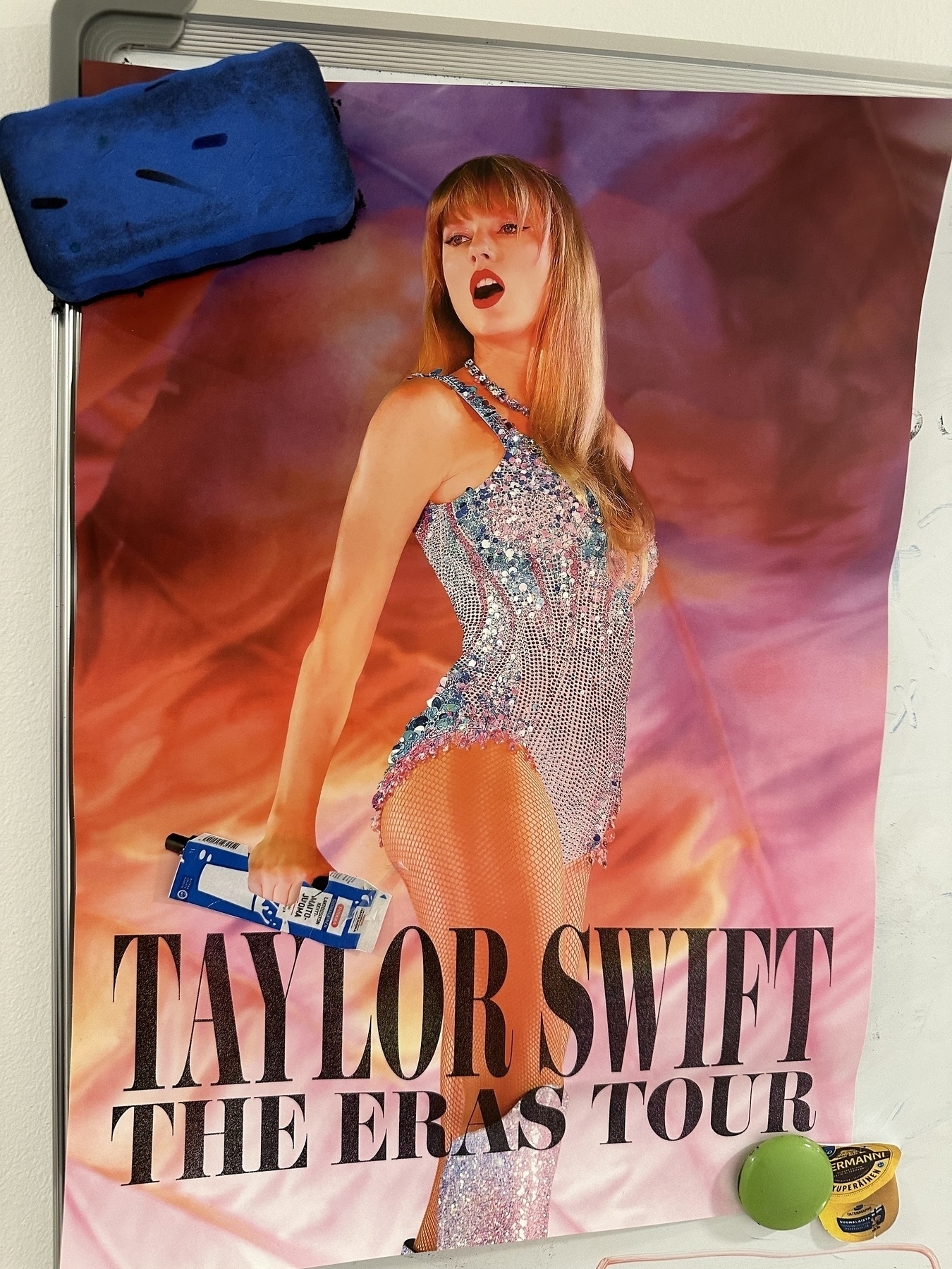 Taylor Swift Eras Tour miniposter on a whiteboard. Taylor looking right over her shoulder and instead of a microphone she has a carton of milk on her had. 