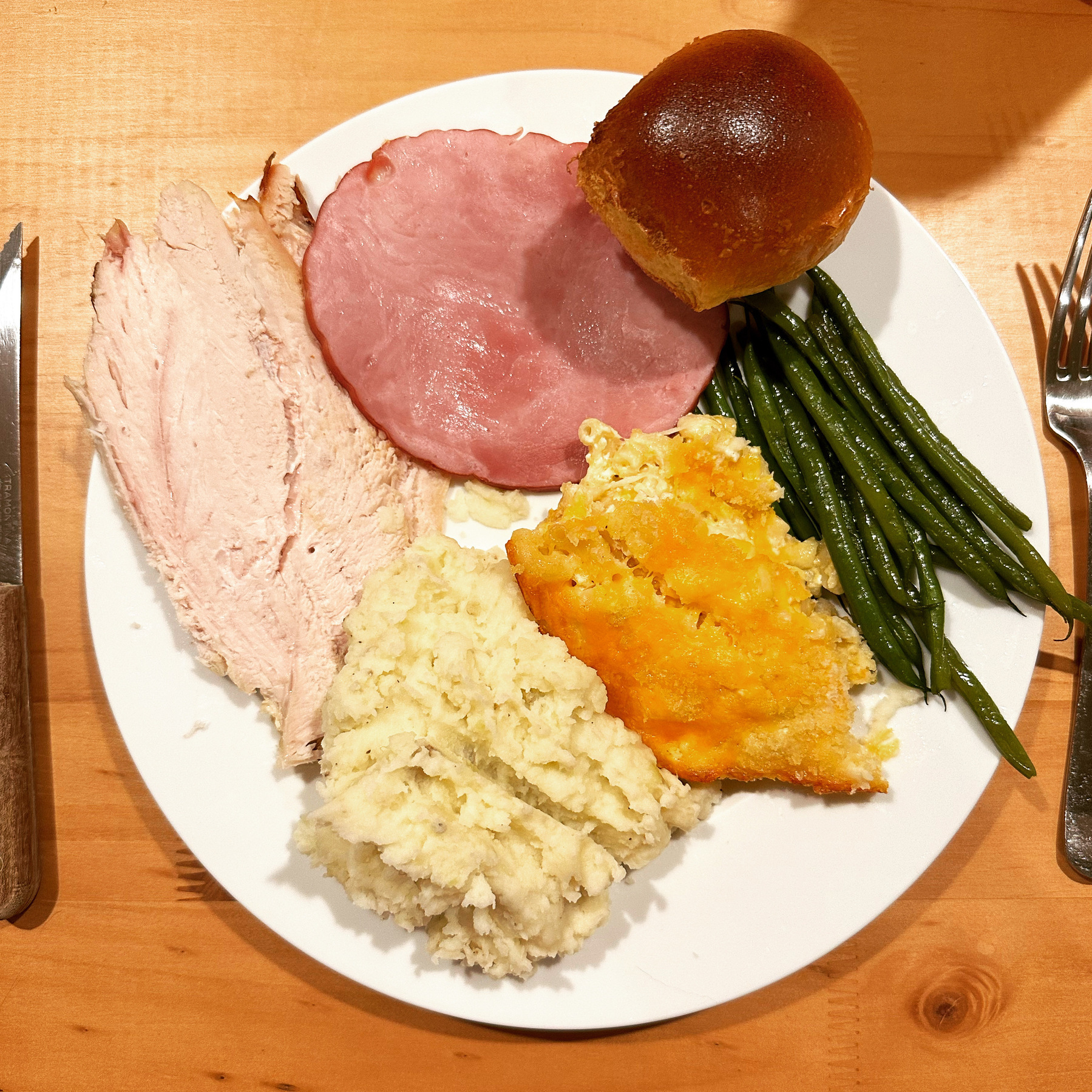 A dinner plate topped with turkey, ham, a roll, green beans, mac and cheese, and mashed potatoes.