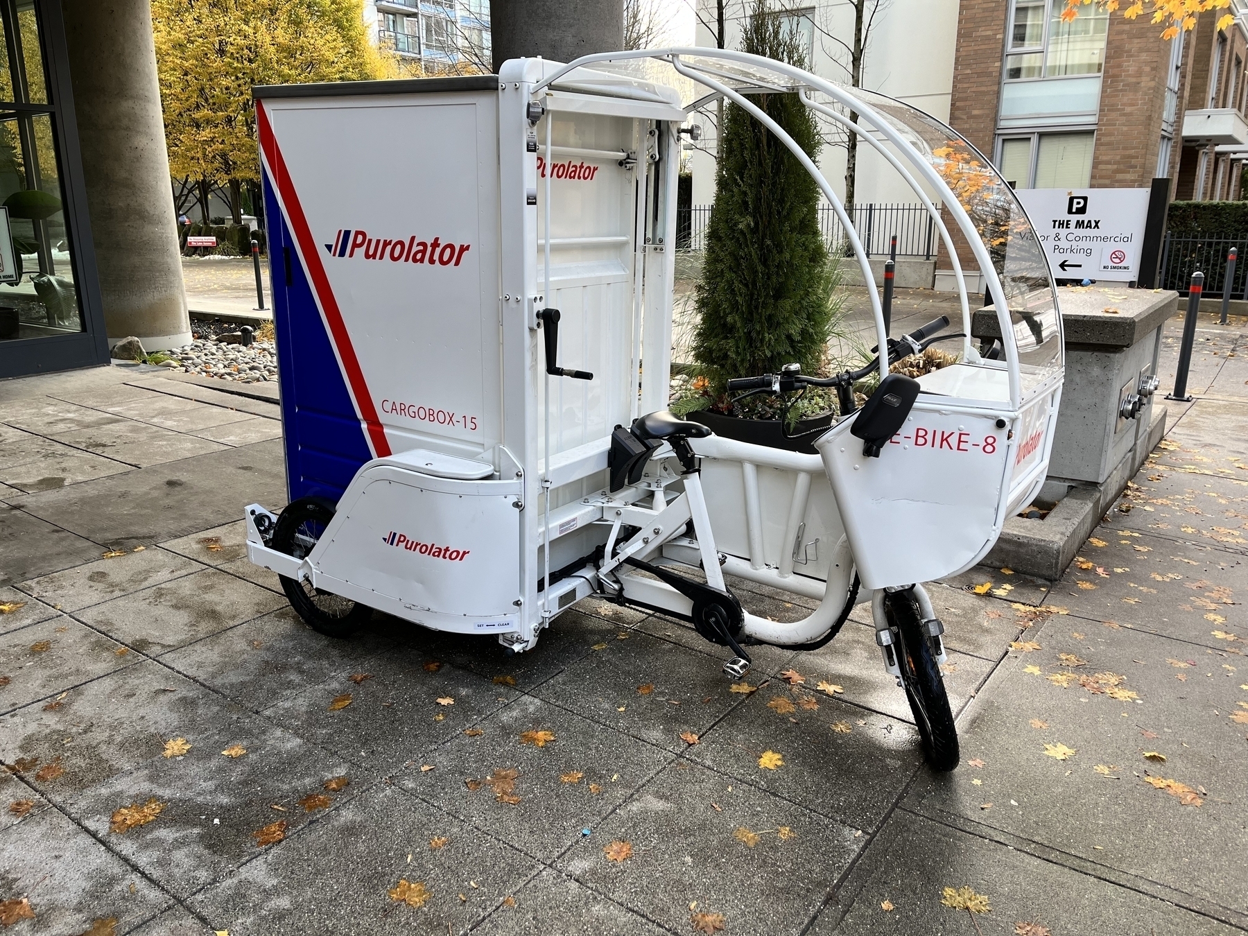 An electric cargo trike with the Purolator logo on it. The bike has a windshield, and a large enclosed rear cargo box that is approximately the size of a refrigerator.