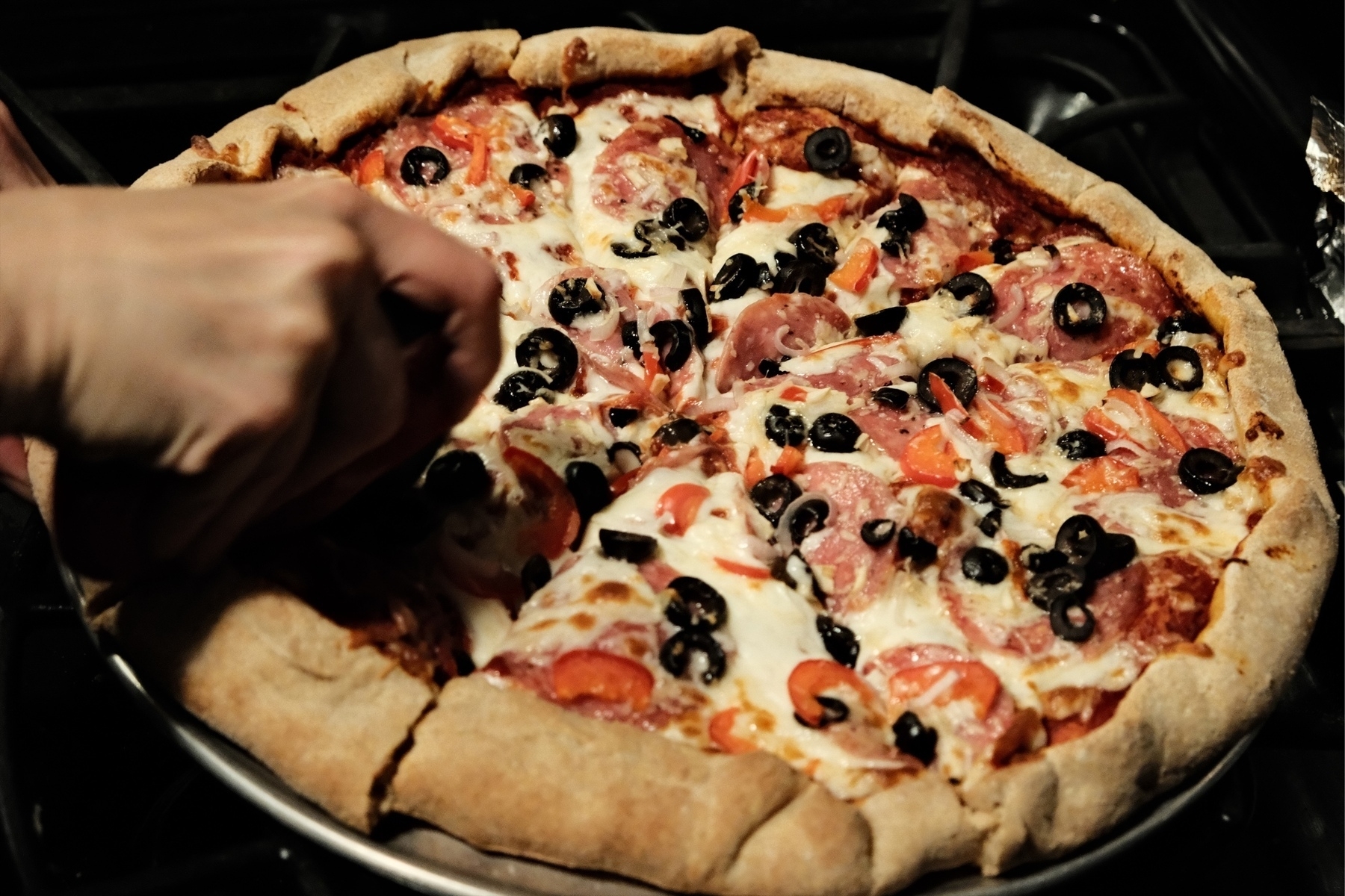 a hand with a pizza cutter slicing a thick crust pizza with salami, shallot, olives, cheese, and tomato sauce.