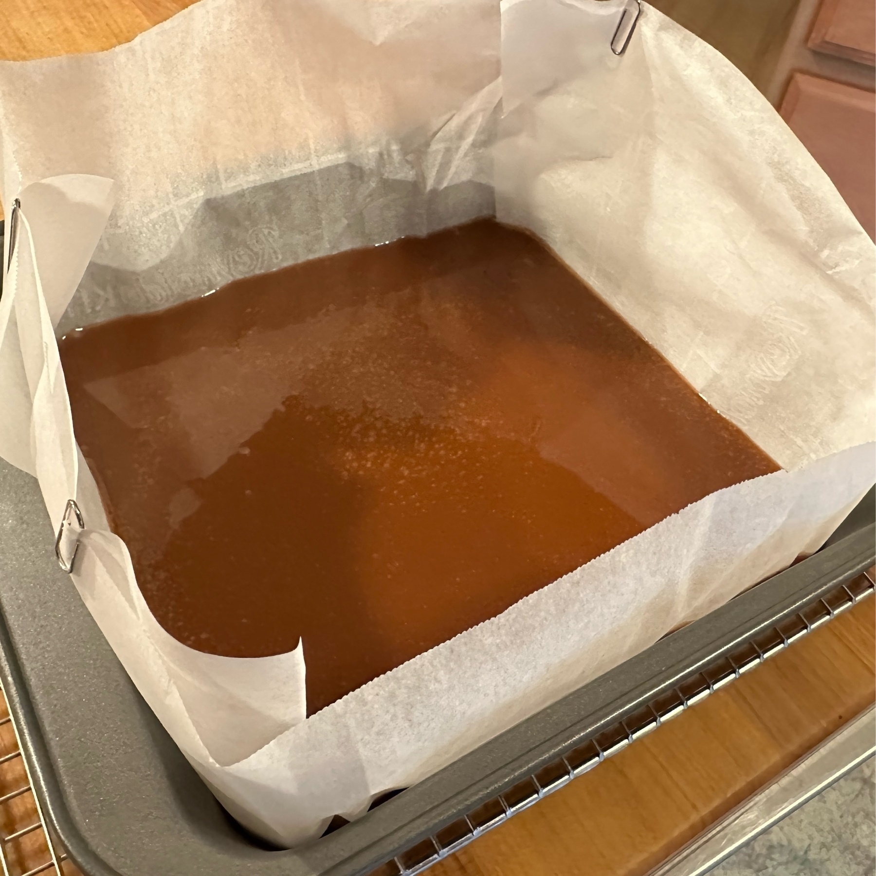 a parchment lined pan of fresh poured caramel. The pan is atop a grate and a cuttingboard.