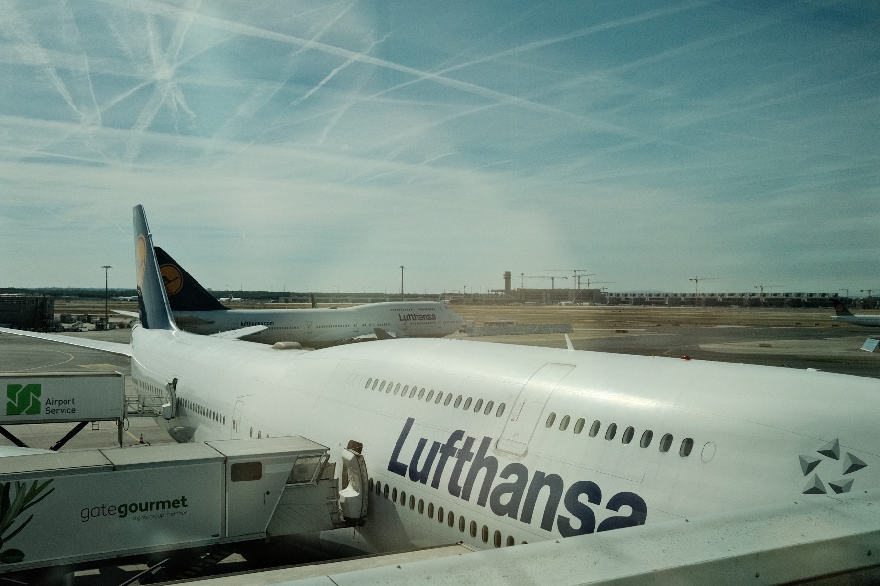 A Lufthansa 747 at the gate and a 747 taxiing behind it. Mulitudes of intersecting contrails mark up the hazy blue sky