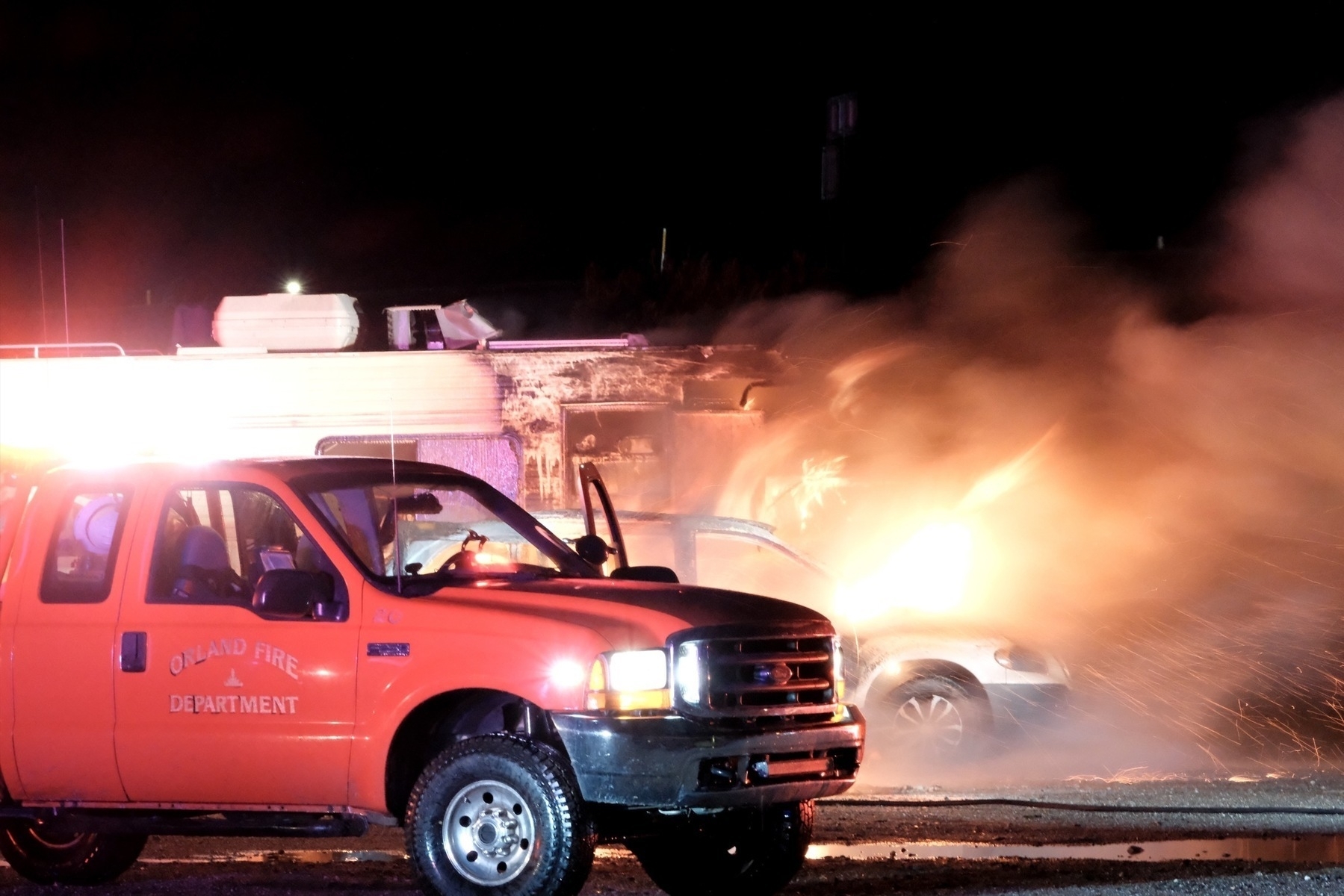 Night time photo of the front half of an RV on fire along with a car parked right next to it. Flames are roaring out th front of both vehicles. An Orland Fire Department truck sits in front of it all.