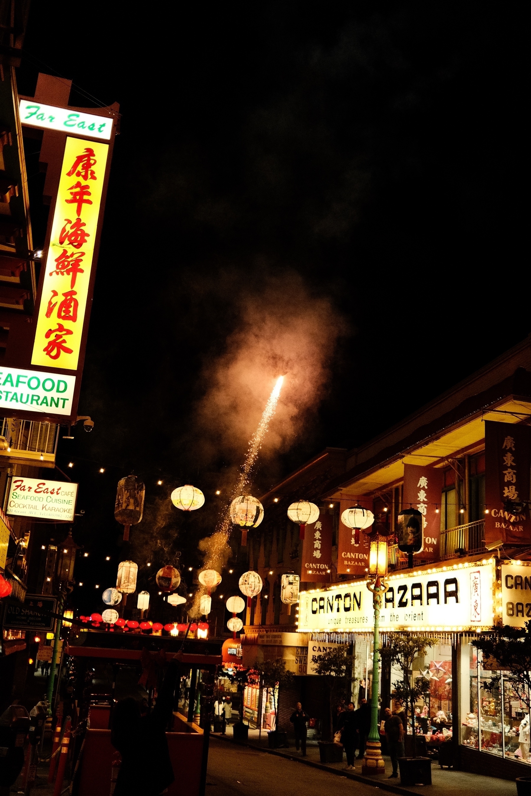 a sparkling bottle rocket shooting anive the dark street with red lanterns suspended above.