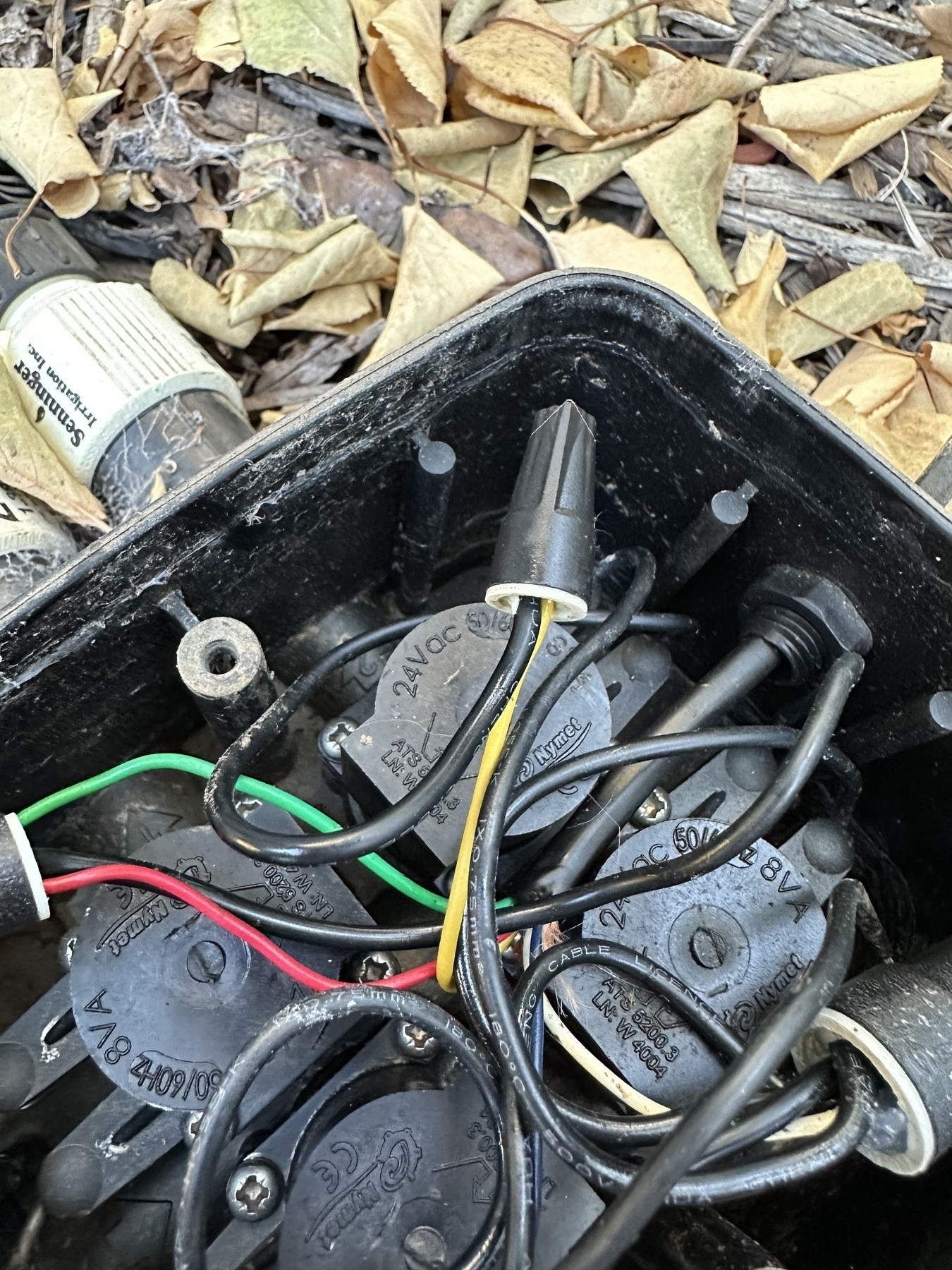 Bundles of 12V irrigation wires in a compact above ground black valve assembly, surrounded by dry leaves. Pressure reducers abs drip adapters are visible.