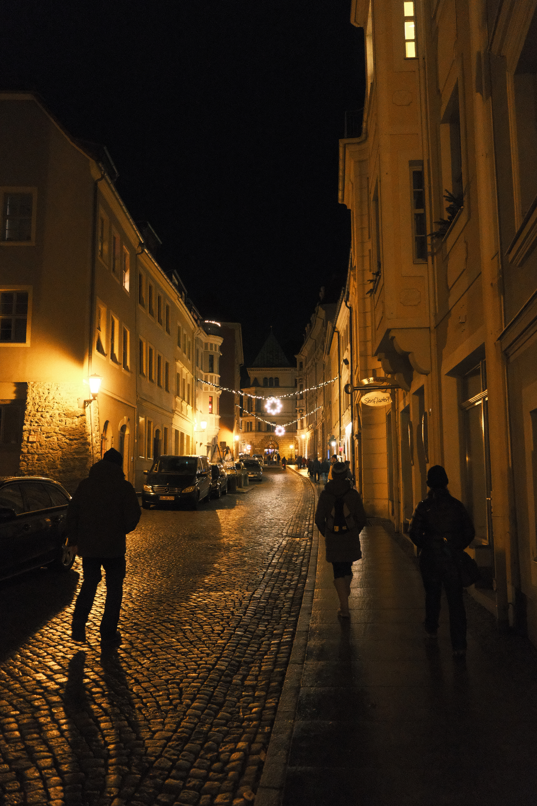 A night scene with warm lights glowing off the cobble streets & three story apartments, with lights strung across the street. Wreaths hang at the center of each light strand. Silhouettes of people with long shadows are walking down the pedestrian friendly street.