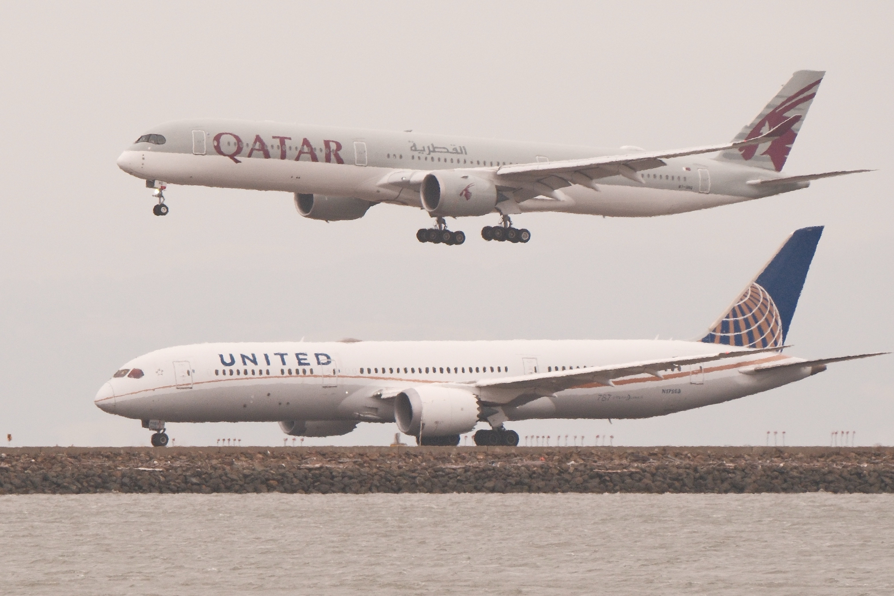 A Qatar A350-1000 lands on SFO's 28R as a United 7879 starts its take-off roll on 28L. The bay is just visible in front of the airport's runway landfill. The A350-1000 has its characteristic expanded uncarriage with six total wheels, while the 787-9 looks sad, feeling left out with only four wheels per main landing gear.