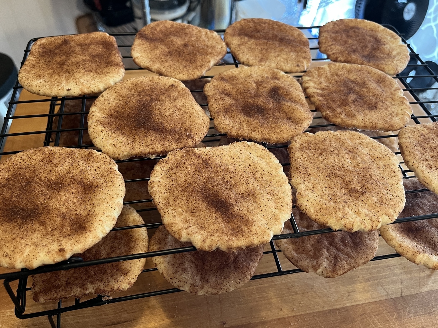 Two stacked cooling racks with well baked, cinnamon covered round & flat browned snickerdoodle cookies. It’s all placed on a wooden cutting board.