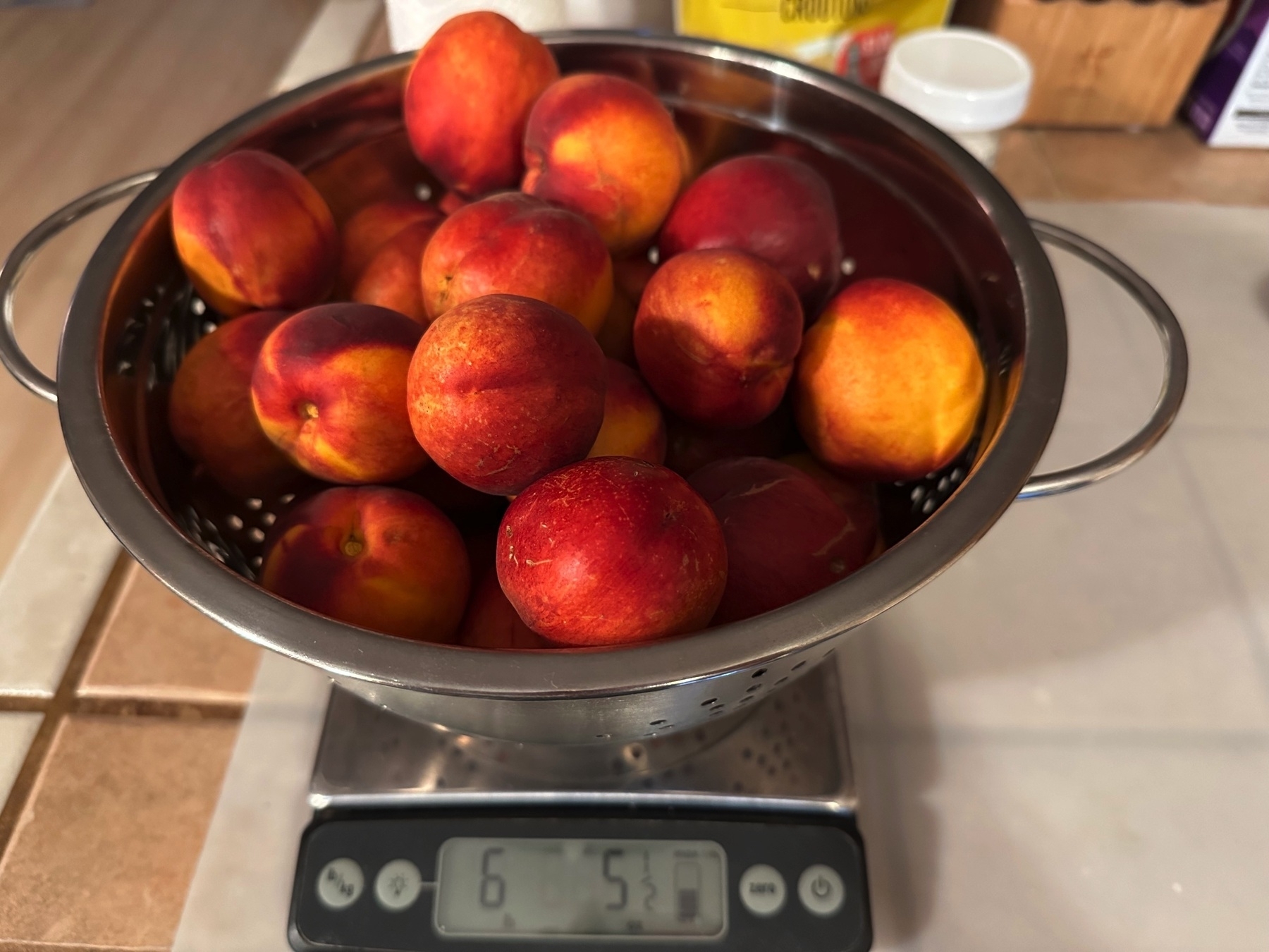 A collander of very ripe dark red to orange colored nectarines sitting on a scale. The scale is on a plastic covering upon a tile counter. The scale reads 6 pounds and 5 ounces, so there’s about 6 pounds of necartines.