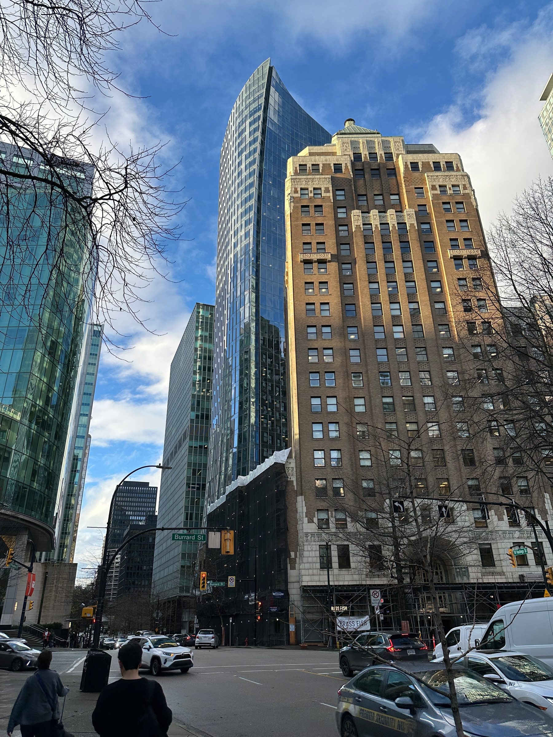 The Marine Building with its glass curves, its top half lit by sun, against a blue sky and partly cloudy skies. The streets are busy and the trees have no leaves. 