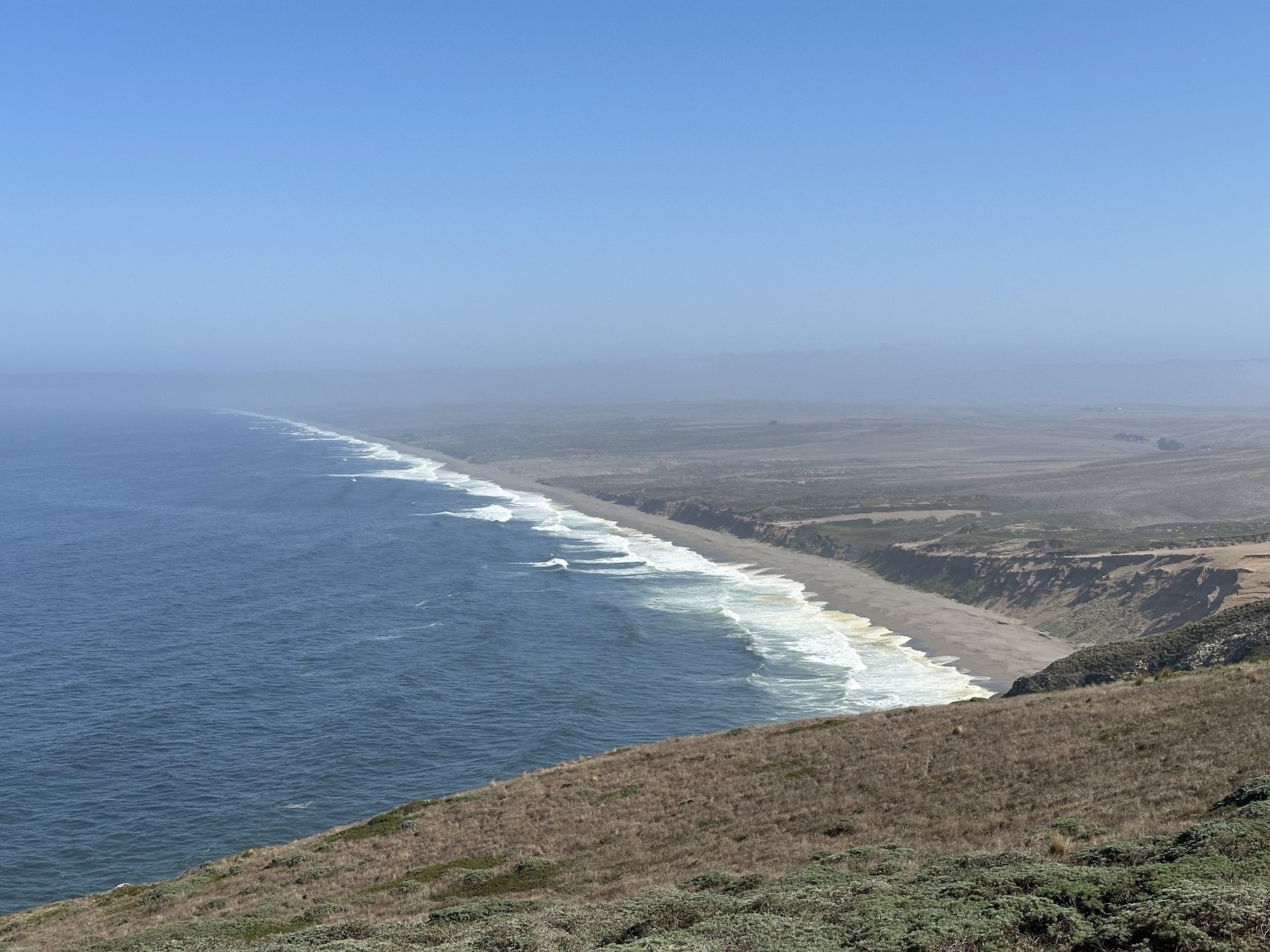 A Long Beach strand faded into the horizon. The surf breaks in white seafoam along the shore. The day is clear, if a bit hazy.