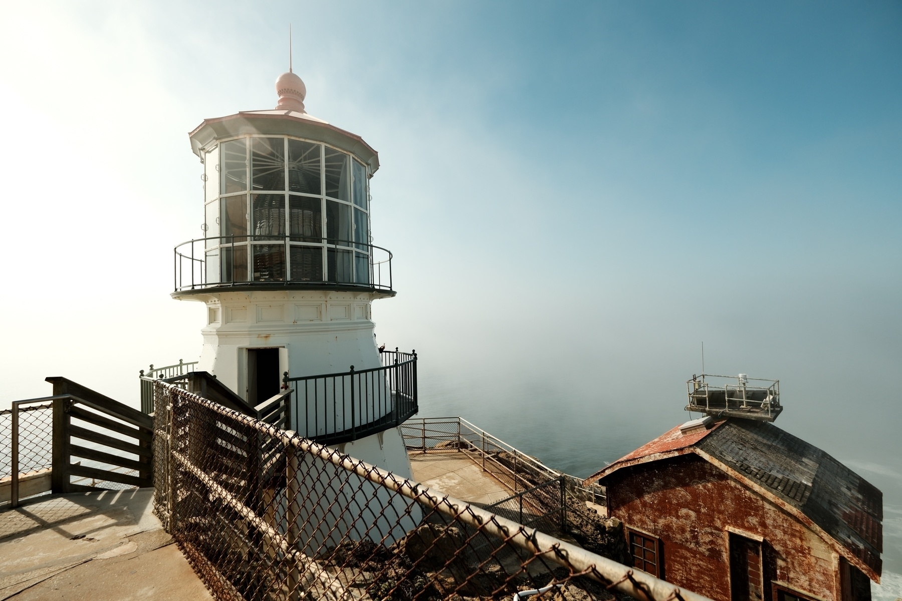 A view of the iconic red fog horn house on the right and the lighthouse on the left. The fog is clearing with a view port of blue sky on the right. The sun illuminates the scene.