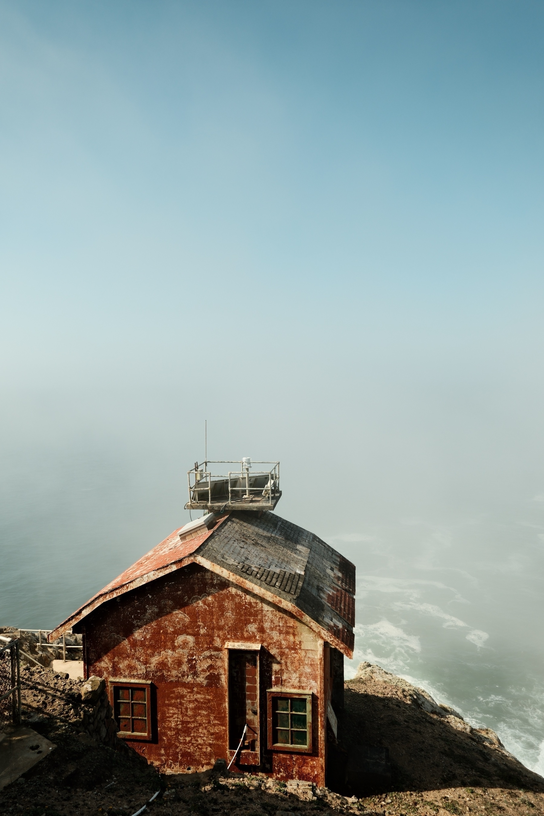 The iconic red fog horn house with a hazy calm ocean behind it. The house is rustic and is in need of a fresh coat of paint. It has a modern electric foghorn and LED beacon setup on the peak of its roof. The building cast a stark shadow to its right (north).