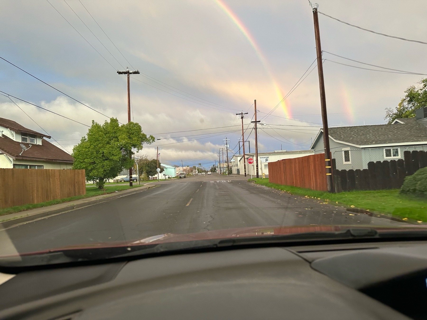 A double rainbow beyond a residential street lined with utility polls, wood fencing, and green ground cover. The main rainbow has its bottom obscured by a cloud & there’s some visible rainfall in the distance.