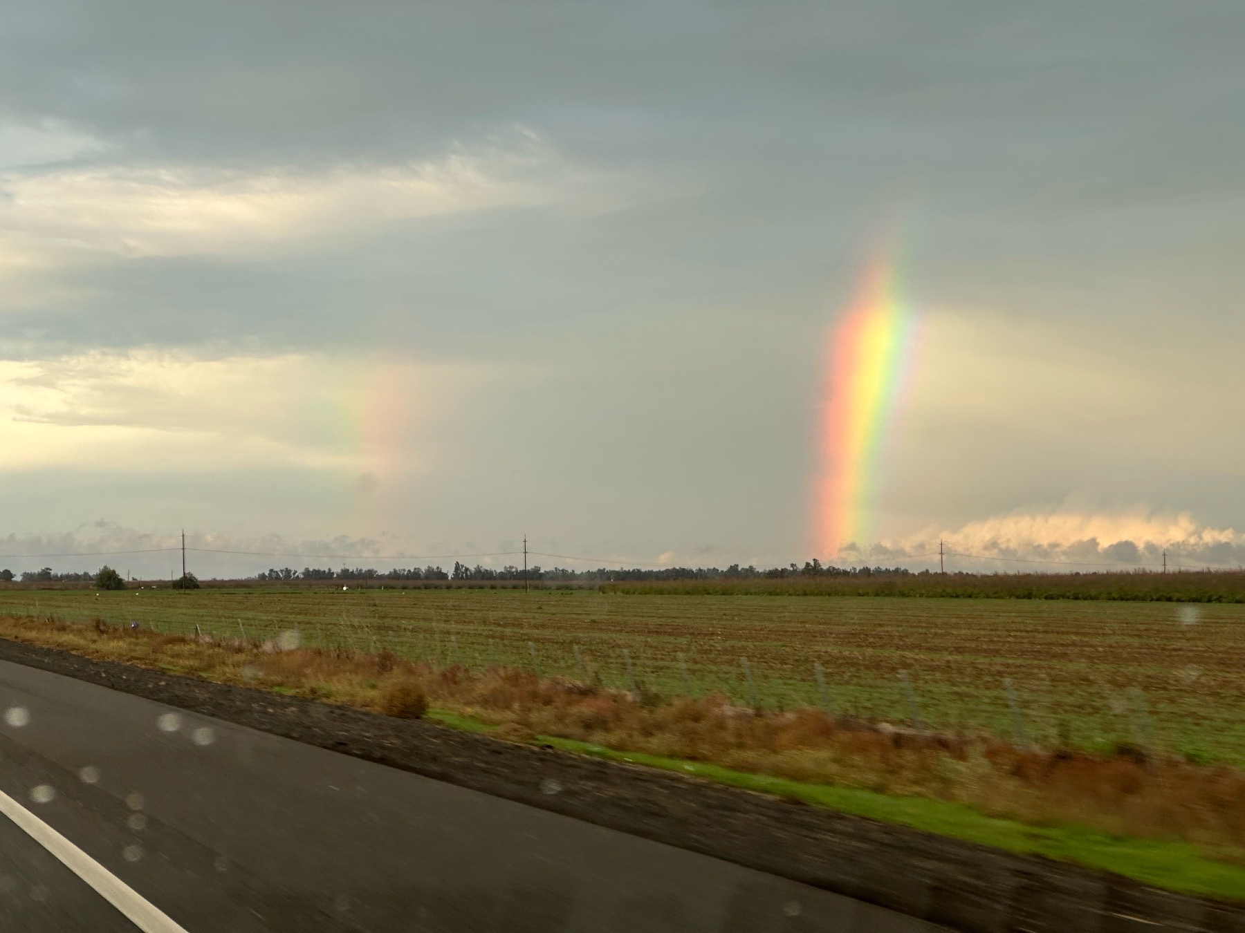 A view out of a car of a double rainbow, including a very bright main rainbow that disappears into stratus above, over a furrowed field. The sun is barely illuminating some lower cloud tops.