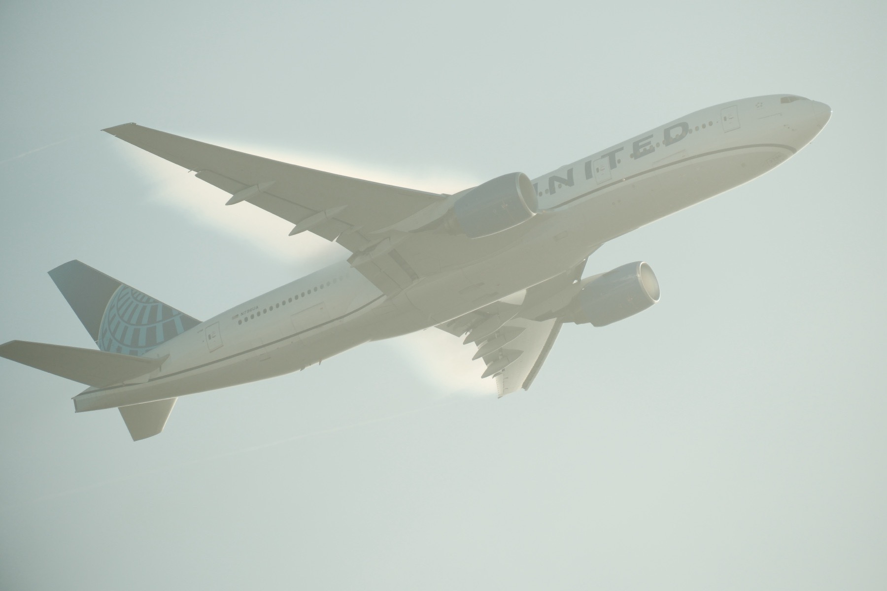 A United 777-200 taking off, gear already up, with slats down and flaps slightly visibly down. The slats are shadowed as the sun is behind the aircraft (though out of frame but causing a lot of glare). Wing condensation is visible above and behind the rear wing edge. There’s also visible condensation in the left engine. The aircraft fills the whole photo. 