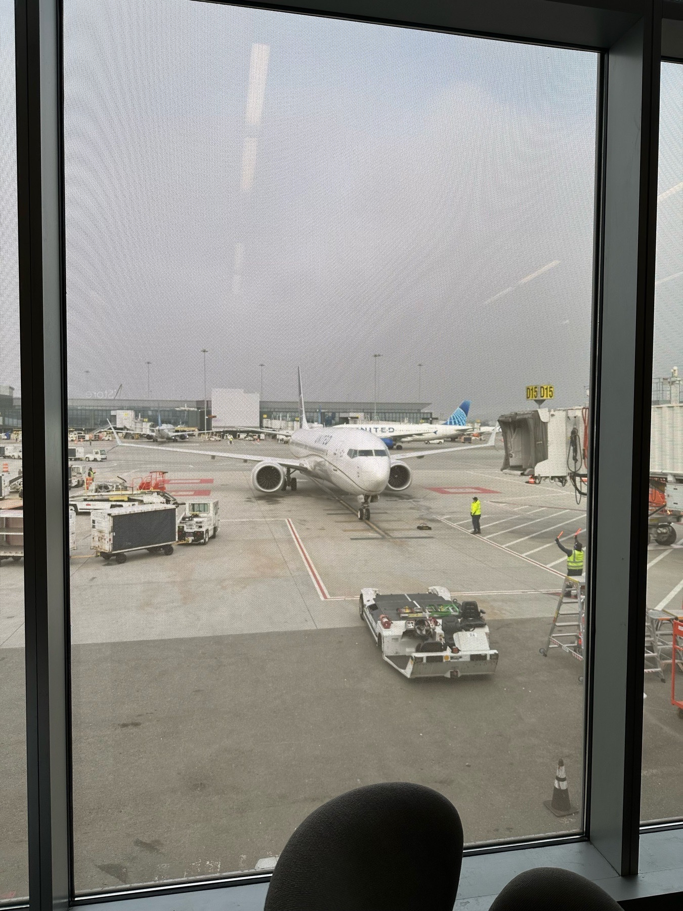 A United 737 Max 9 noses into gate D15 in front of an aviation lover’s stippled glass nightmare. Equipment cars surround the aircraft. It’s a foggy day 