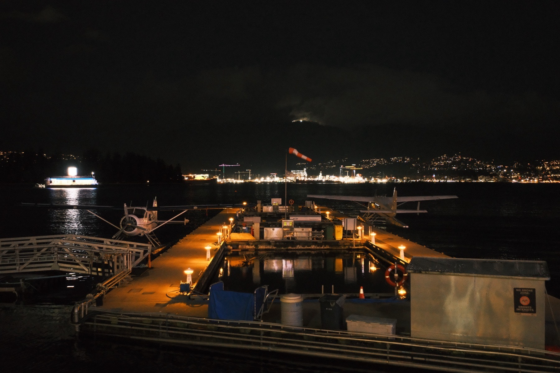 A night view of two single prop seaplanes, opposite sides of the photo, at their own warmly lit docks. A windsock is flying at a slight angle toward the ground. There’s a strange cloud shrouded light above the horizon. Across the harbor are hills and lights. Just beyond the docks to the left, there’s a floating chevron gas station.
