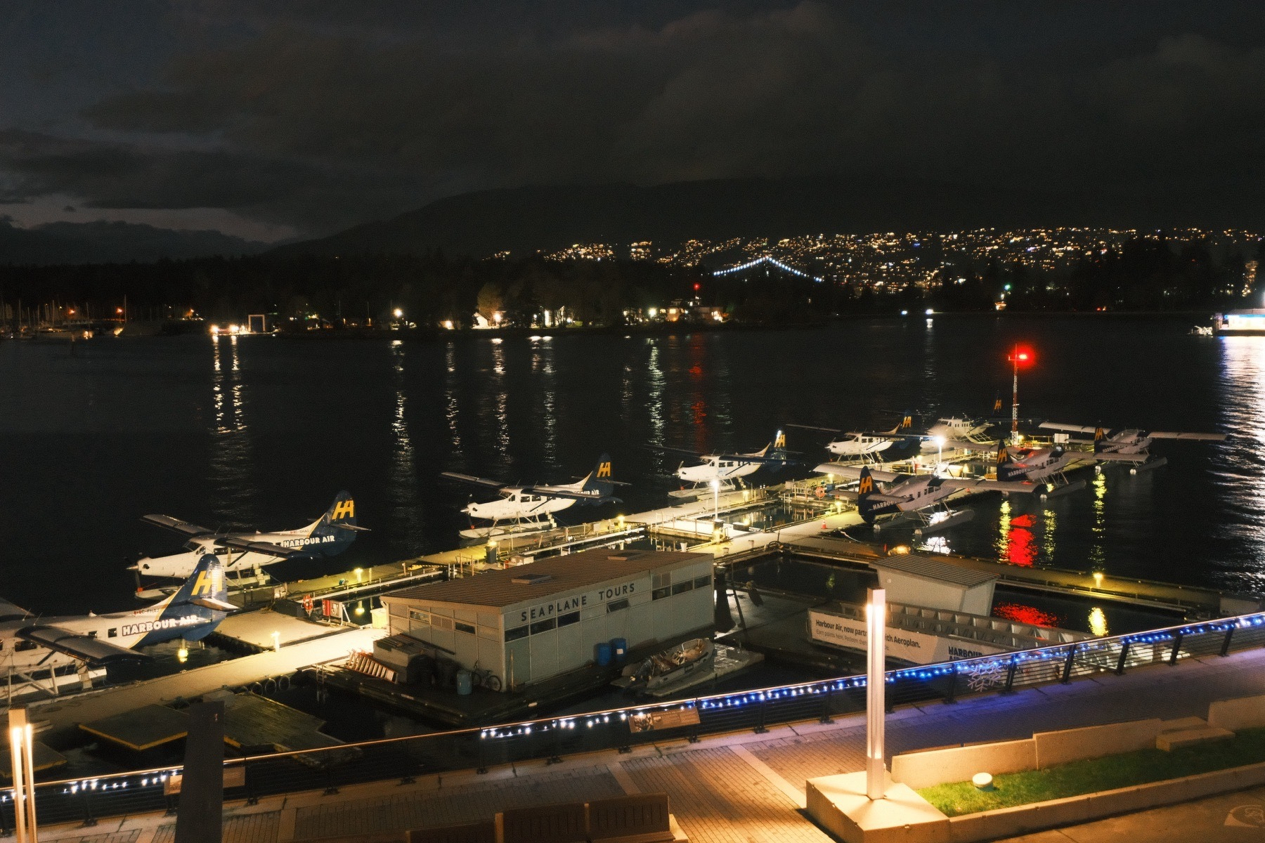 A nighttime photo of five brightly lit single prop seaplanes all lined up in a column along their brightly lit dock. A bright red beacon is at the end of the dock on the right. In the distance, across the harbor, are hills surrounded by artificial lights. A short span of suspension cable lights (including one tower) of the Lions Gate bridge is visible.