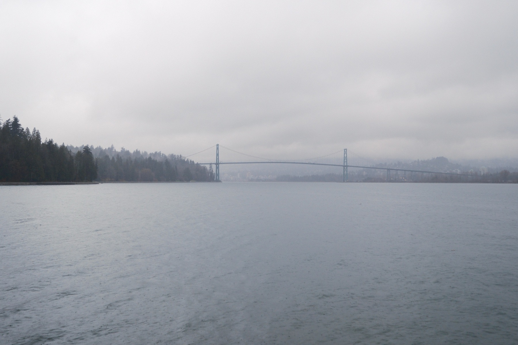 Lions Gate Bridge in the distance, at the horizon. Vancouver Harbor is calm and the moisture haze fades points of land before the bridge. Low stratus clouds everywhere.