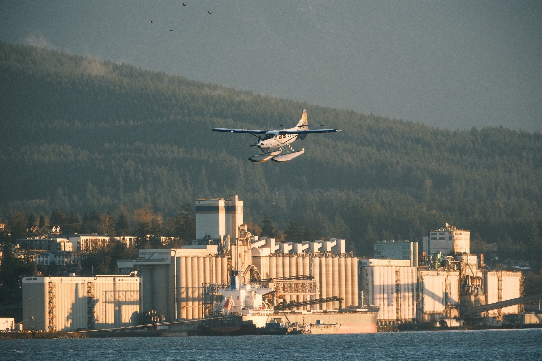A seaplane descends above the harbor, with the sun reflecting off its body. A large industrial facility with series of tall tanks looms in the background . The hills behind it all are forested with a few wispy clouds.