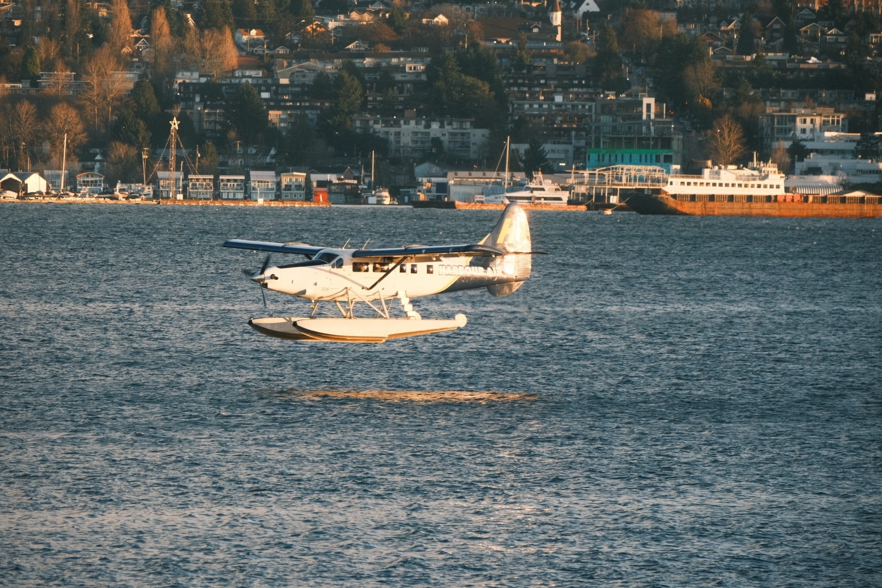 A seaplane flies a few feet about harbor waters as it lands, its airframe reflects in the harbor waters. The morning sun lights up a background of ships, port facilities, trees, and buildings built up a gentle hill.