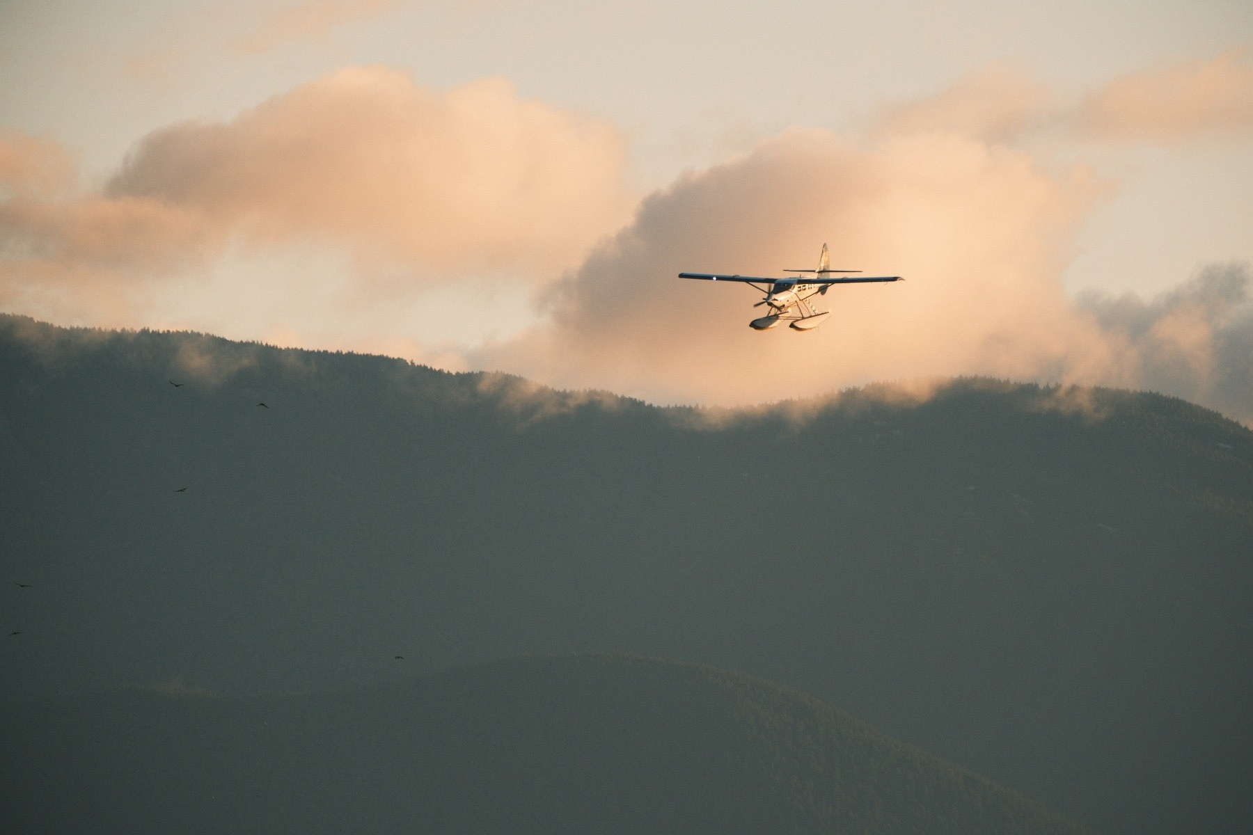 A float plane descends in front of fluffy light orange clouds and unlit mountains below