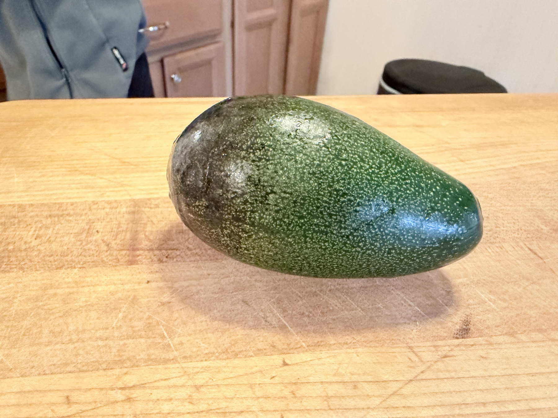 A "bacon" variety avocado on a cutting board. The bottom quarter has turned dark green compared to the rest of the skin. Its skin is densely spreckled.