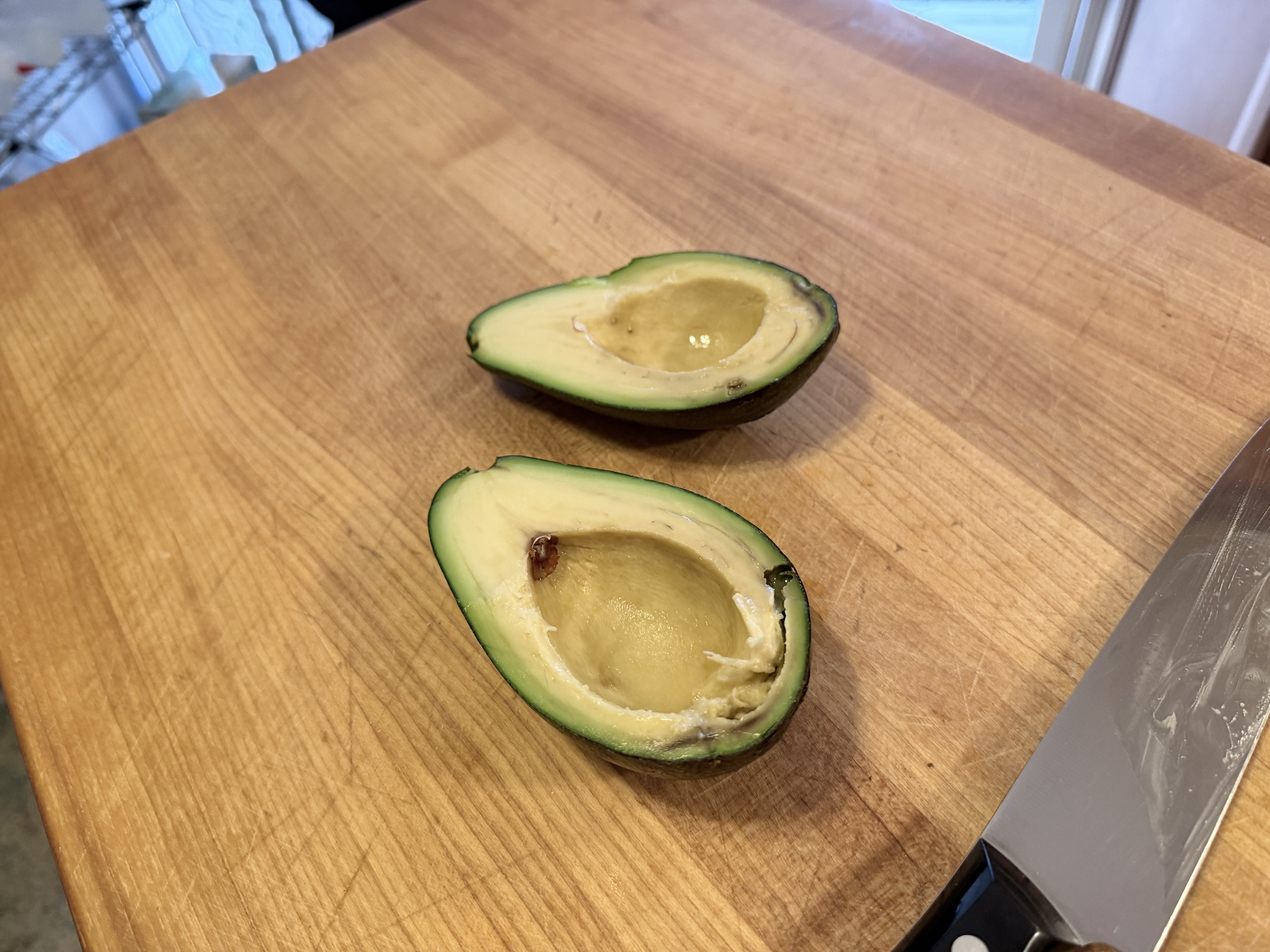 The "bacon" avocado is cut in half, and pitted, showing where the puncture happened on the bottom, and that some insect(s) likely already dug in for a meal. Its flesh looks soft and is greener near the skin and a olive-yellow toward the pit. A knife lays to the left of the fruit.