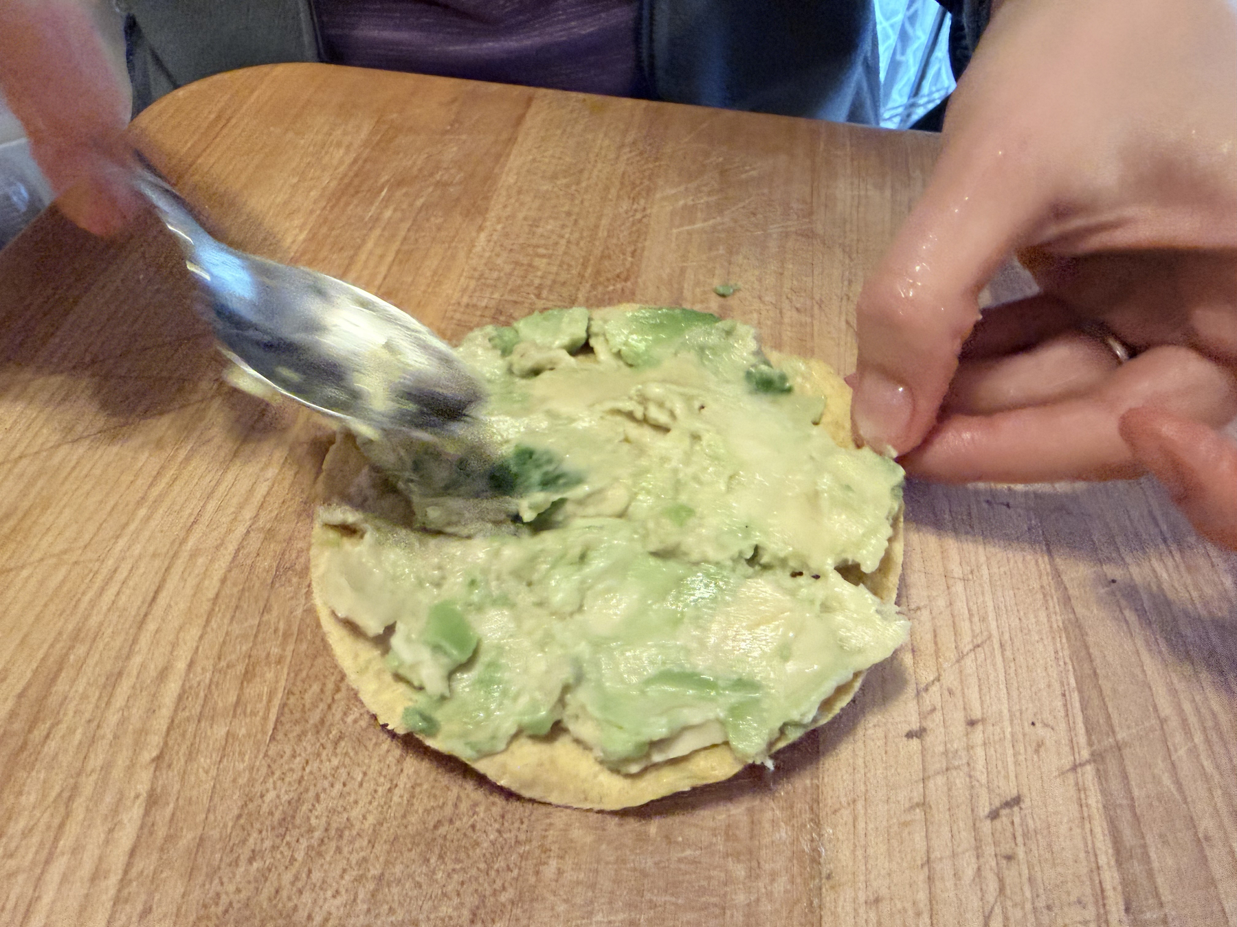 On a cutting board, the "bacon" avocado is being smeared across a tostada shell. It looks very creamy and indeed it was easy to spread. One hand is holding the tostado and another is smearing the avocado flesh with a fast moving spoon.