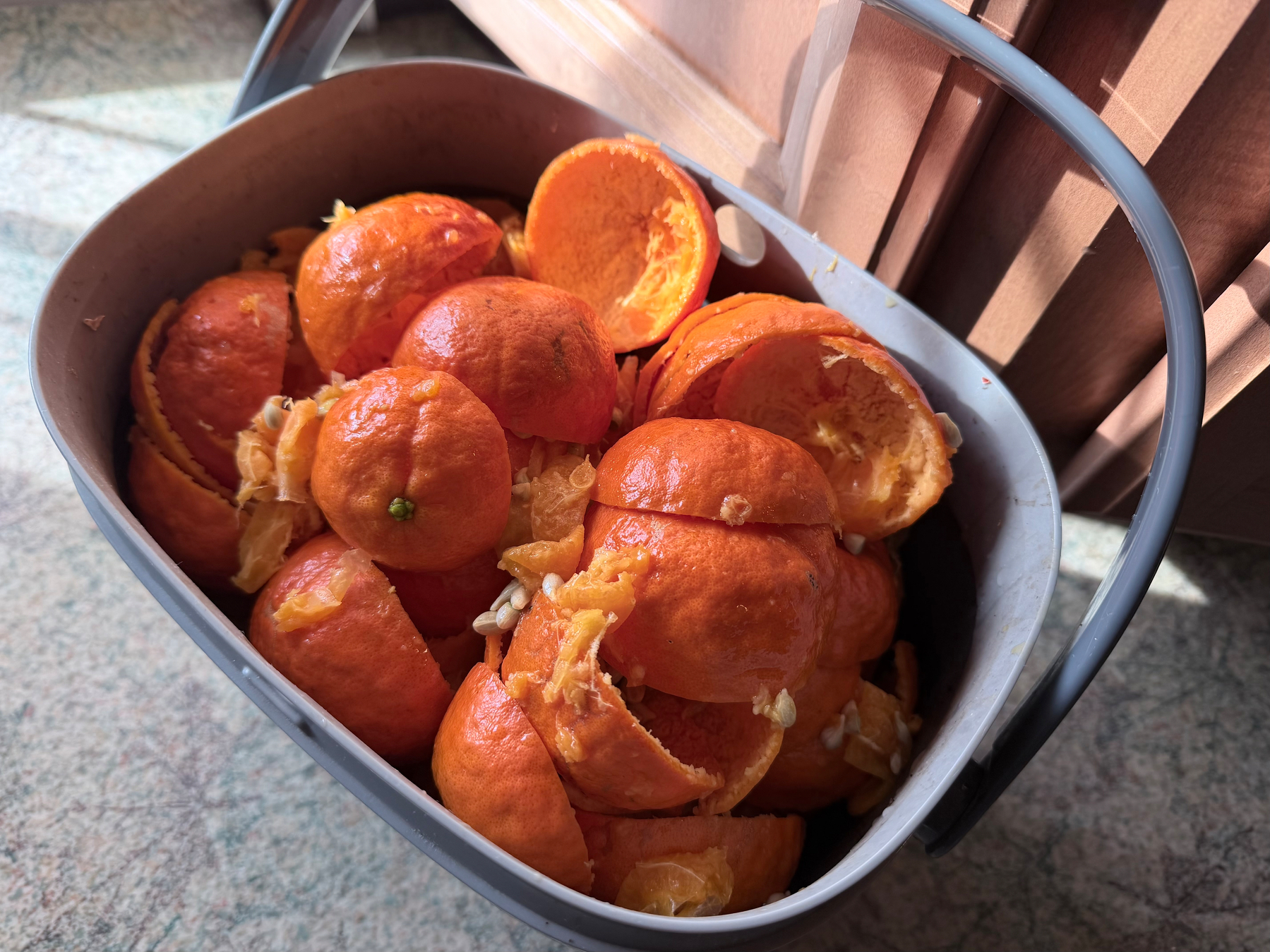 A small 1/4 gallon (maybe?) plastic bucket full of bright orange mandarin skins, pith, seeds, and pulp. It’s sitting in front of a sliding glass door, against cabinets on a linoleum floor with a speckled leaf pattern.