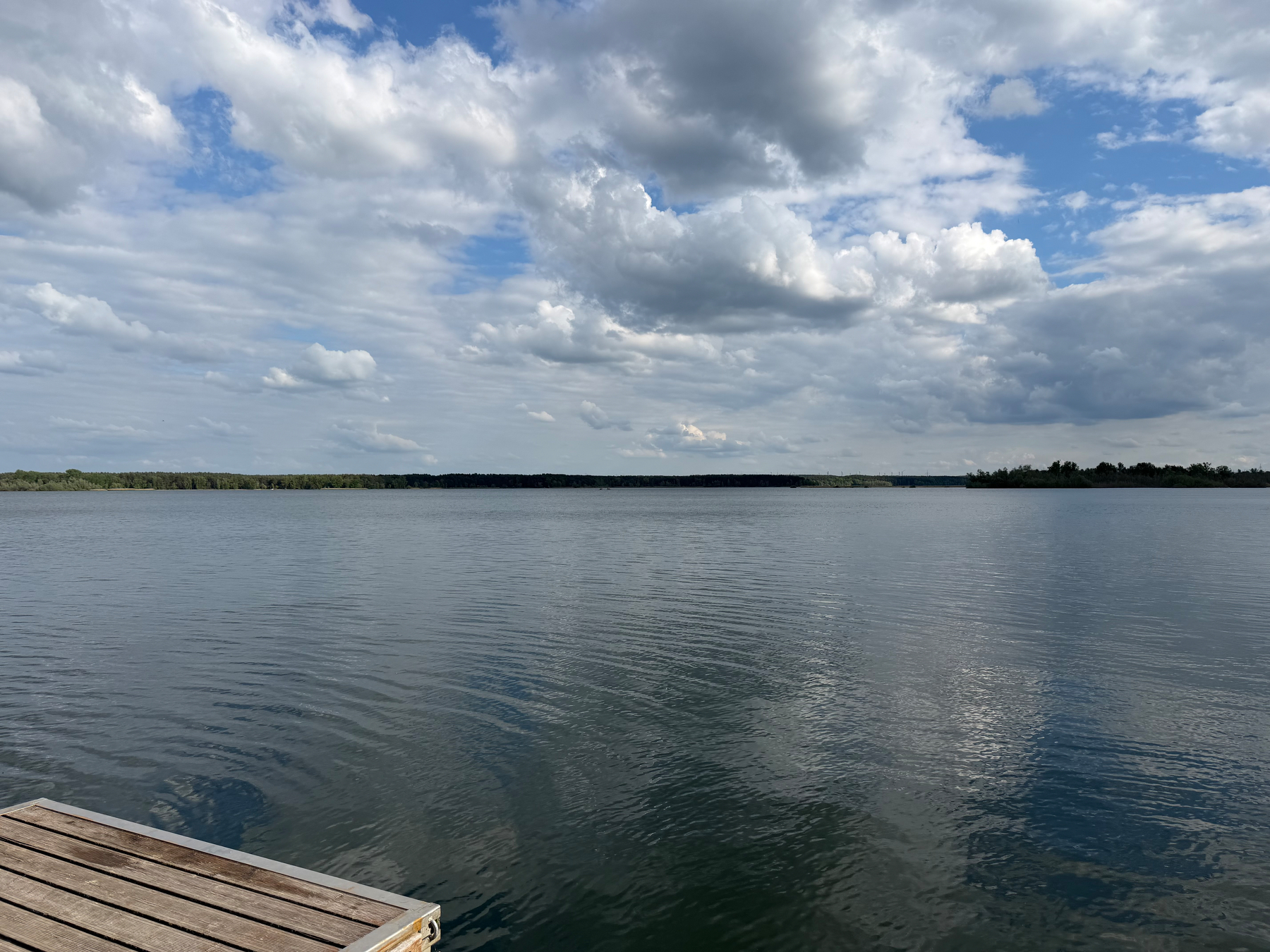 Clouds over Talsperre Spremberg, man-made lake in Germany as a result of coal mining. The clouds are fluffy and reflect a little in calm but rippled lake waters. The end of a wooden dock peaks out in the left bottom corner.