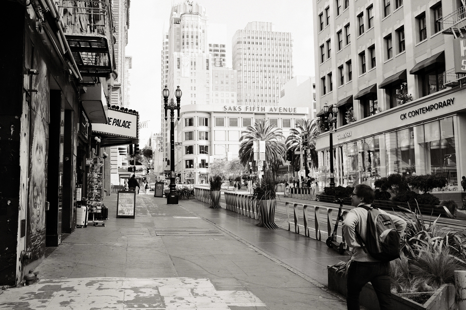 A sidewalk view of Powell and Geary streets, with a person looking and smiling at a building-side mural of a family of monkey or lemurs. Various buildings loom with Saks Fifth Avenue, Swarovski, and CK Contemporary signs. Two classic palm trees sit between the Saks building and Swarovski, likely at Union Square.