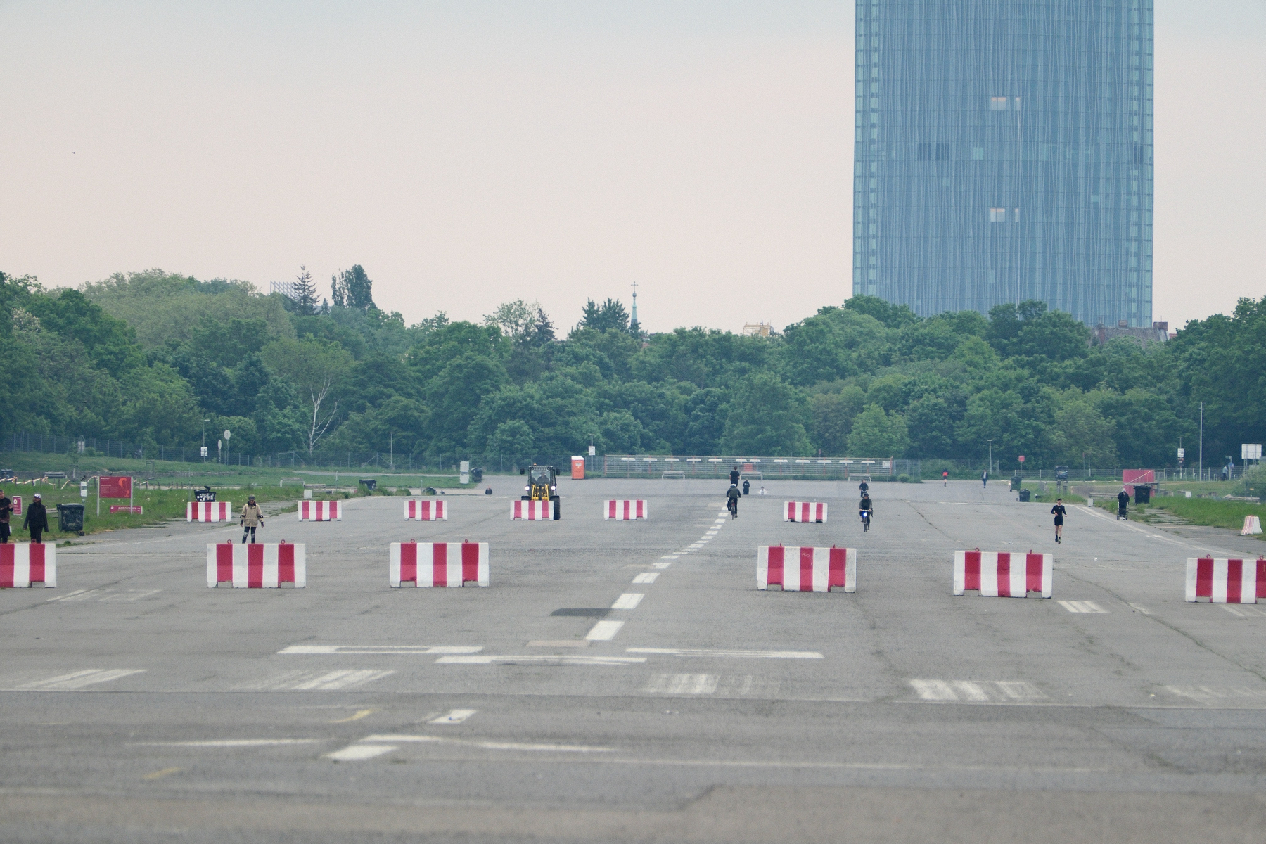 DSCF0059.JPG A view down the right runway of the long-closed Tempelhofer Field in Berlin. Striking K-rails with red and white stripes are placed in rows on the runway with wide separations. There are only a handful of people biking, scootering, walking, and running on the runway. All vegetation is green and in the distance are lots of trees. On the right, a lone sky scraper juts out of the ground from behind the trees.