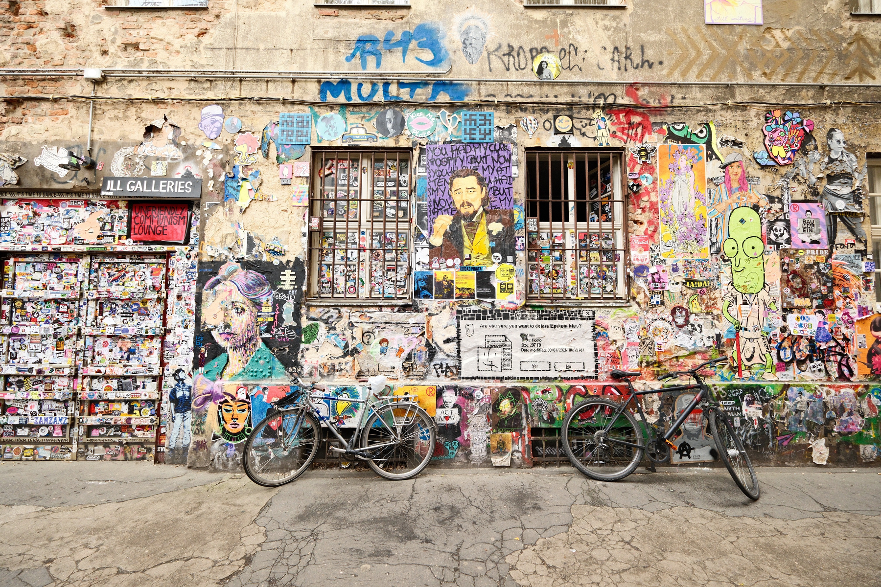 DSCF0161.JPG A building wall with a mixture of colorful art, graffiti, and stickers. There's one prominent piece of art prompting to delete the Epstein files. Two bikes are parked in front of the building wall.