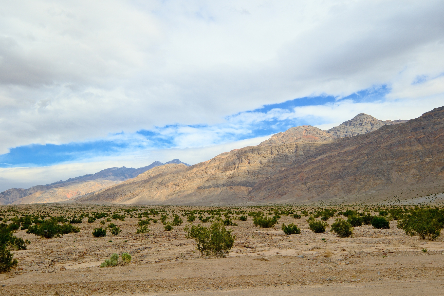 DSCF5466.jpg Overcast skies, with a ribbon of blue breaking through, let some sun break through to highlight rugged mountains, brown with patche sof red, that obliquely frame the photo. Dark green shrubs, maybe mesquite, dot the landscape from near the photographer and into the distance. Some of the closer mountains have layers of dark and light rock. Those closer mountains are shadowed by clouds.