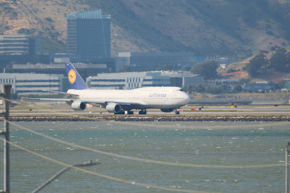A Boeing 747, owned and operated by Lufthansa, taxies at SFO to 28L. Its at an angle, but the sun is fully illuminating its right side. It is set against buildings and brown hills of South San Francisco. The bay in front is a bit choppy and the photo was taken somewhere above powerlines, as they are visible at the bottom edge of the photo.