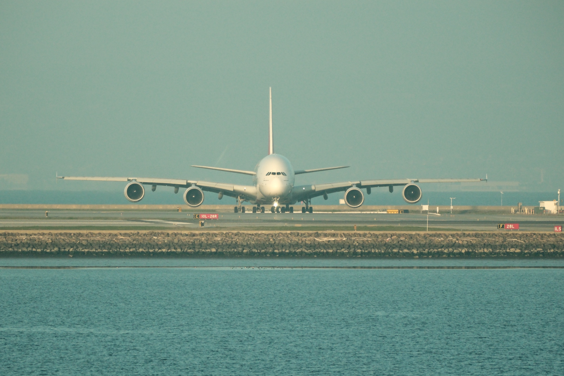An Airbus 380 is nose-on to the camera, showings its clearly thick tail reaching high above the ground. Its nose gear is turning so it is about to leave the taxiway for a runway. The sky is hazy yet still tinted blue. The bay in front of the runways is calm.