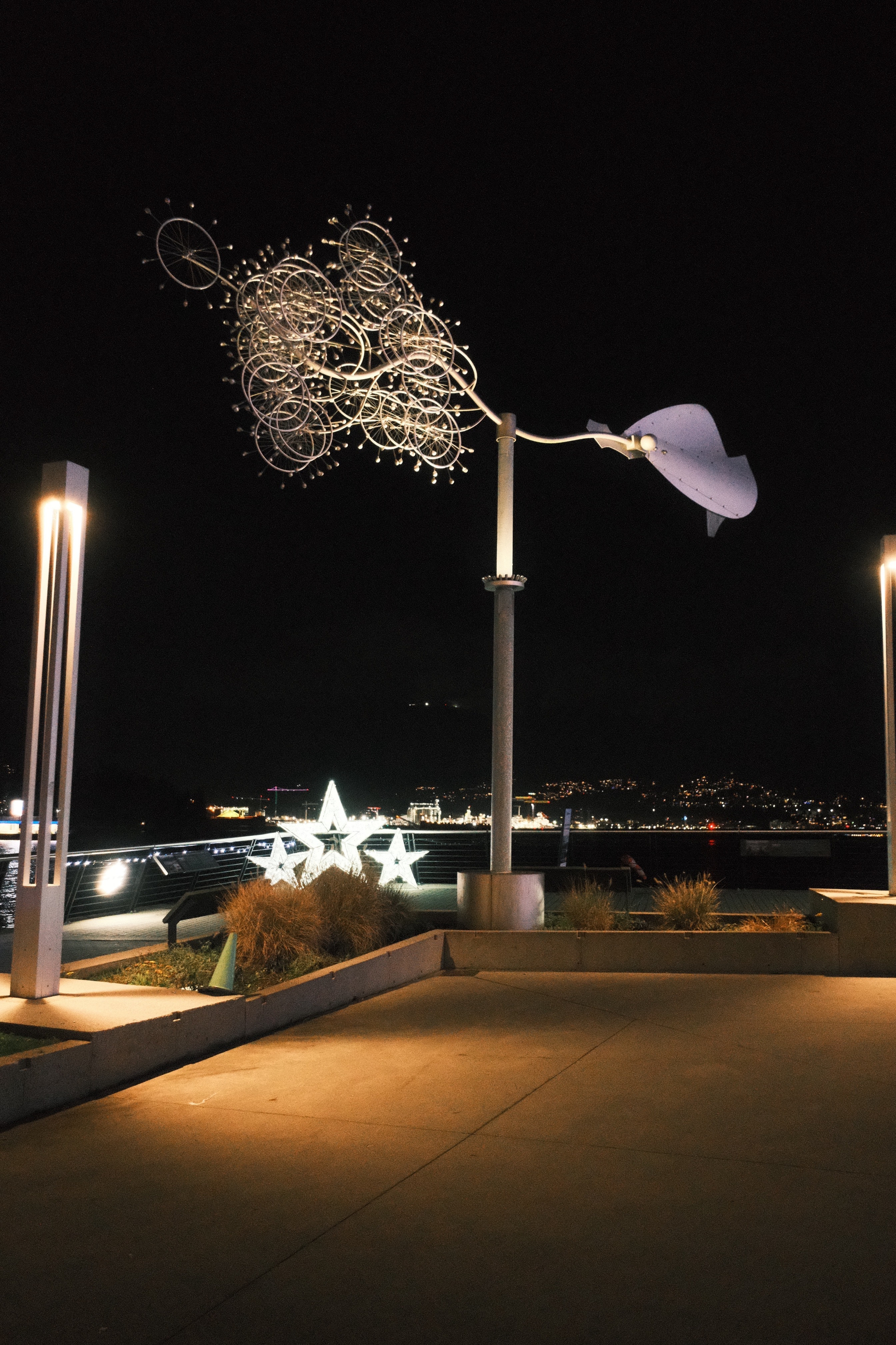 At night, an artistic weather vain with one end as metal outlines of clocks or maybe wheels and spokes. The other end looks like a couple of paddles. It's all lit up with purpose. It does spin but no motion can be seen from the photo. Behind it are three lit up stars, probably christmas decorations for the waterfront. Lights across the harbor can be seen.