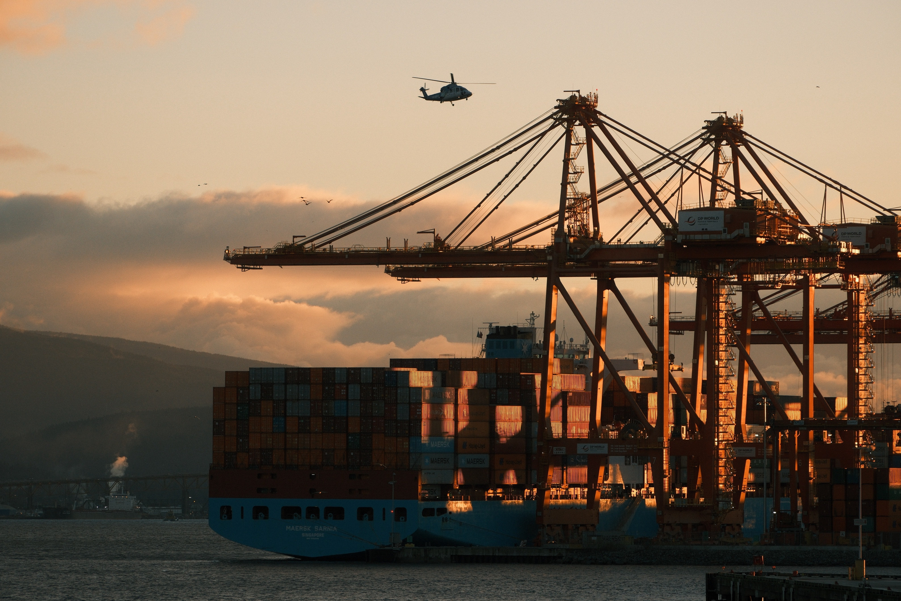 A sunrise lit container ship, with its east-facing side lit up and the rest in shadows. There are clouds looming on the mountainous horizon over Vancouver habor waters, including a group of birds flying in front of the clouds. Large cranes are also illuminated from one side. A helicopter is just above the cranes, approaching a nearby landing pad.