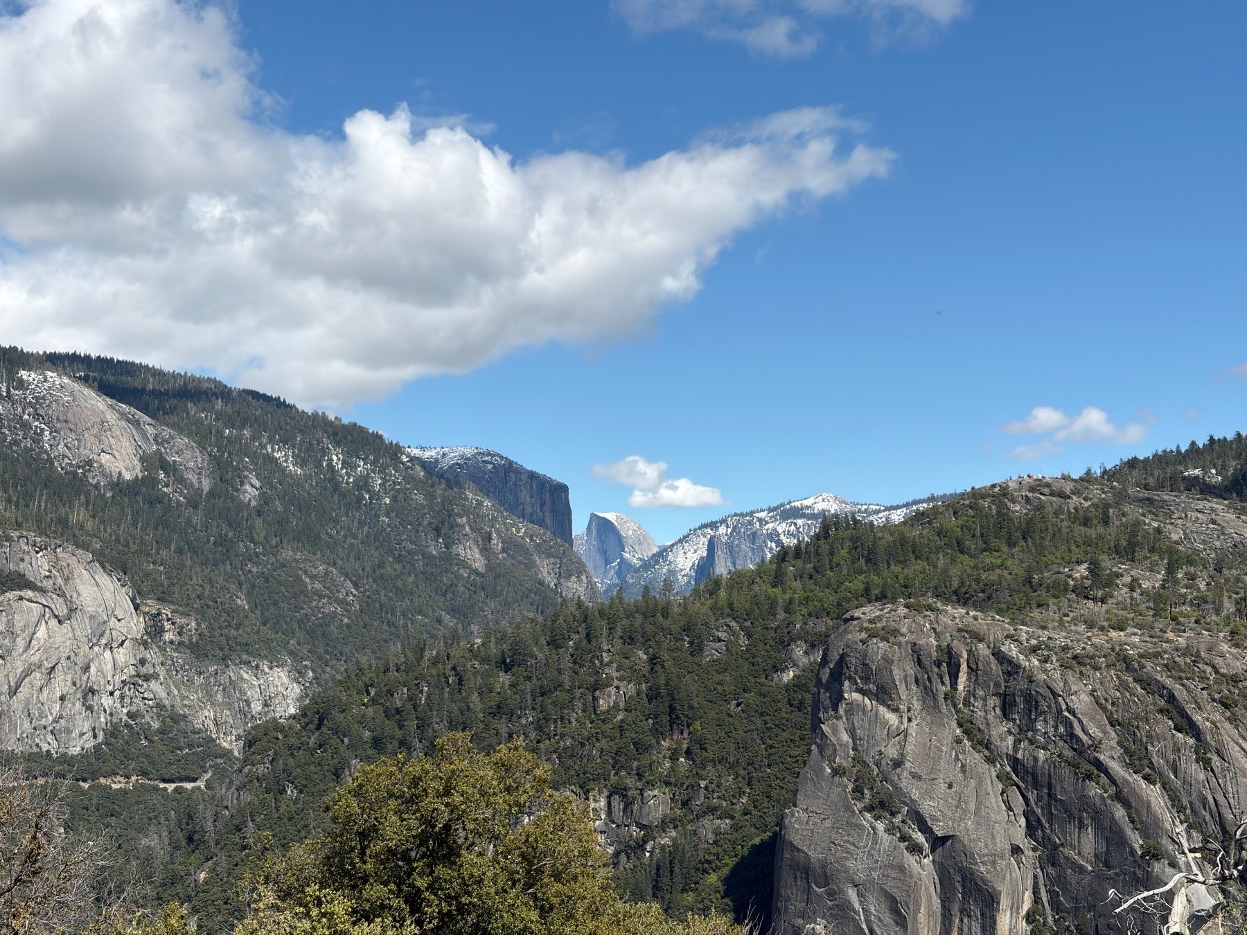 A view down the Yosemite Valley with Half Dome in the center. It’s a partly cloudy day and there’s a dusting of snow atop the mountain peaks. Patches of bare granodiorite contrast with the surrounding conifer forests.