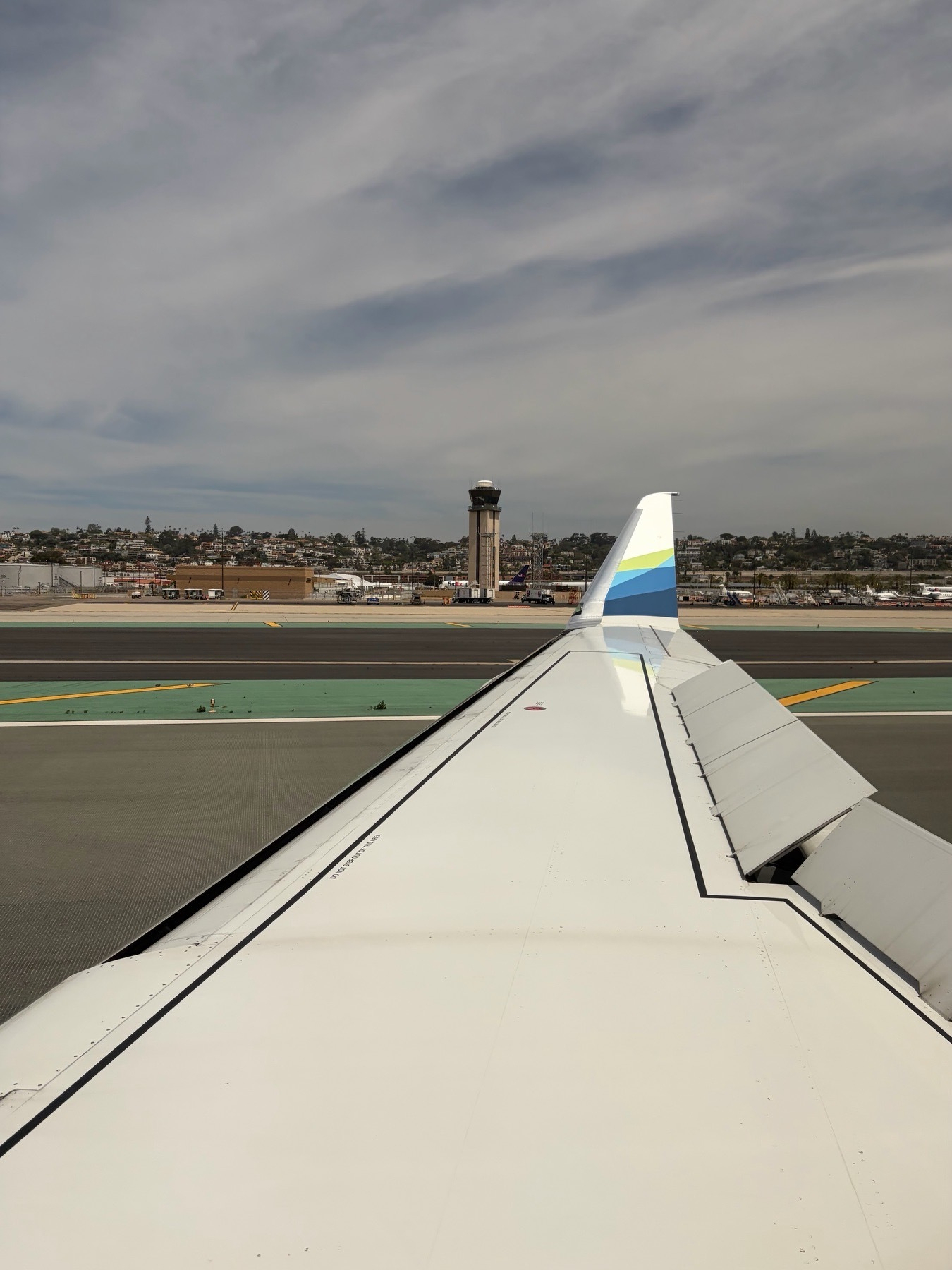 An over wing view on an Alaskan Airlines (SkyWest) Embraer 175 upon landing at San Diego International Airport. The speed brakes are up and the winglet with blue to green swooshes is prominent. The tower is visible along with an assortment of jets, including FedEx. It’s a cloudy day with very thin stratus. 